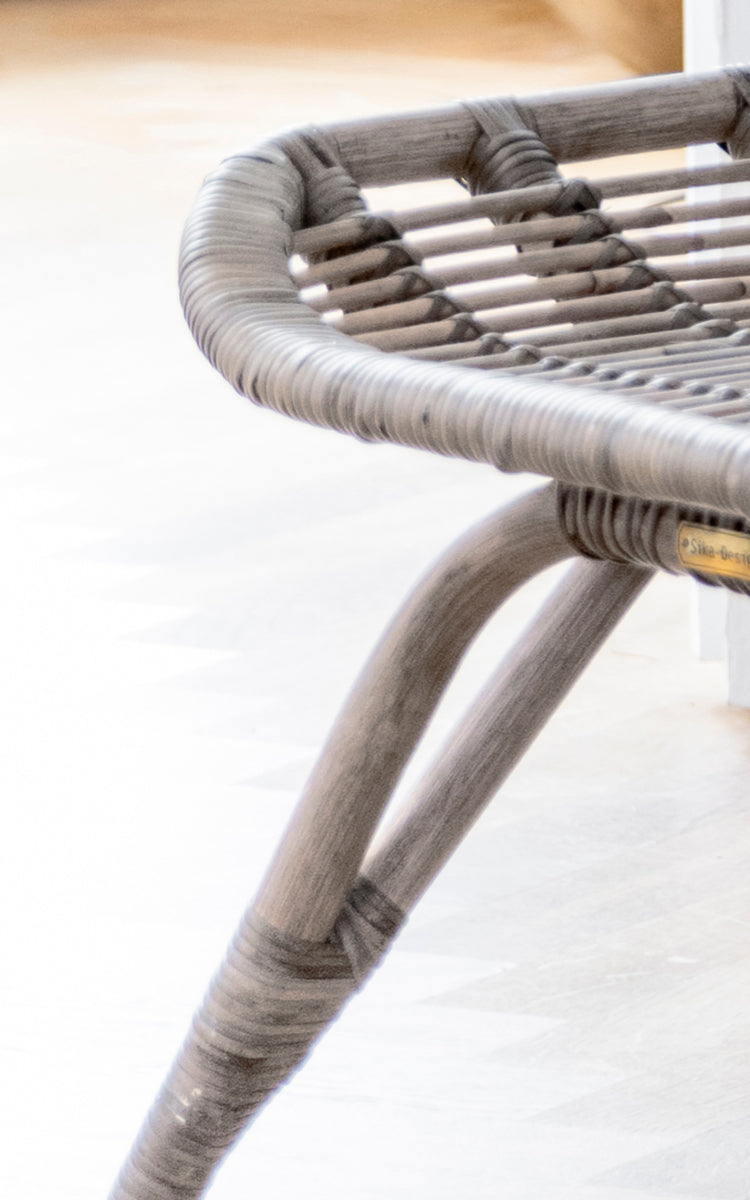 This is a close-up image of Monet Footstool in taupe rattan. Detail of the woven seat top and curved leg frame in soft taupe finish.