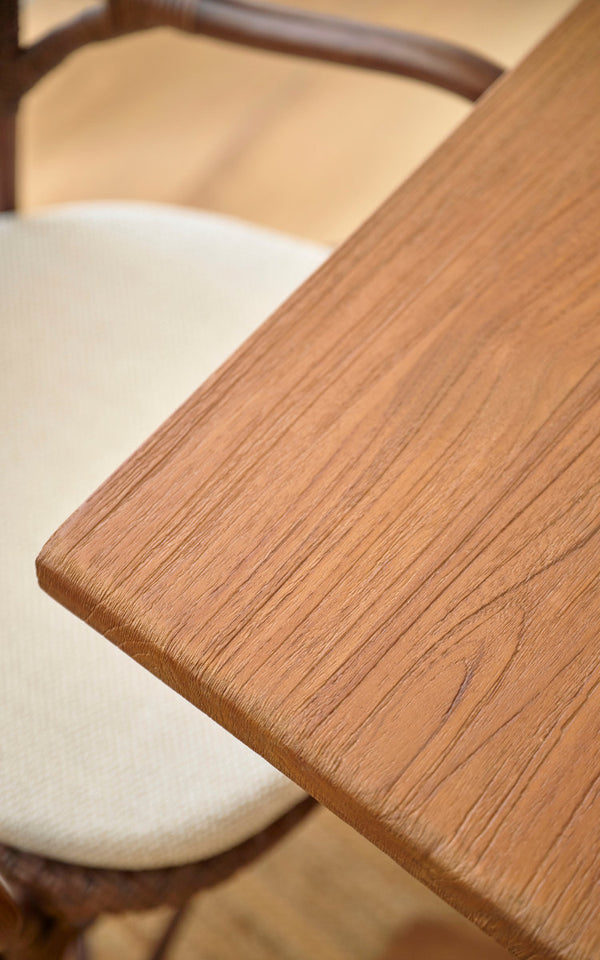 This is a close-up image of Hugo Teak Dining Table 140x90 cm in teak indoor. Smooth teak tabletop edge beside a rattan chair.