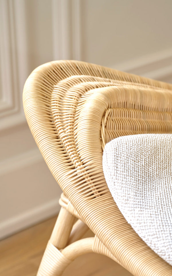 This is a close-up image of Pacifique Lounge Chair in natural core weave rattan frame. Woven shell with white cushion by a paneled wall.