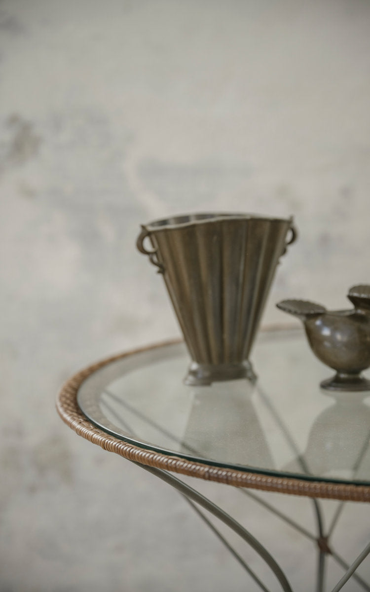 This is a close-up image of Funky Café Table in hazelnut rattan iron frame. Glass top with pewter vase and jug.