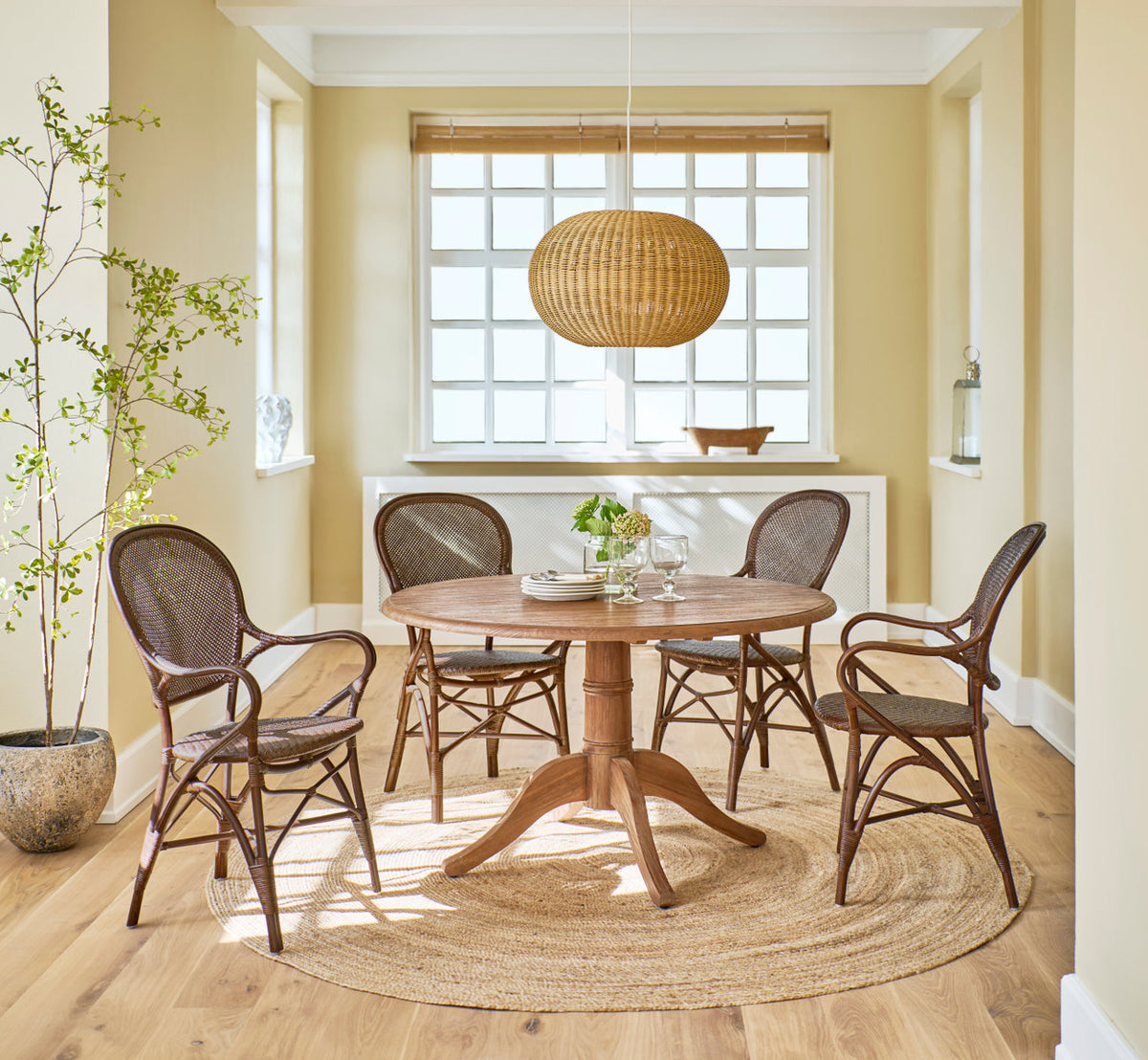 This is a lifestyle image of Rossini Dining Armchair in brown rattan. Round teak table, rattan pendant and jute rug in a sunny dining room.