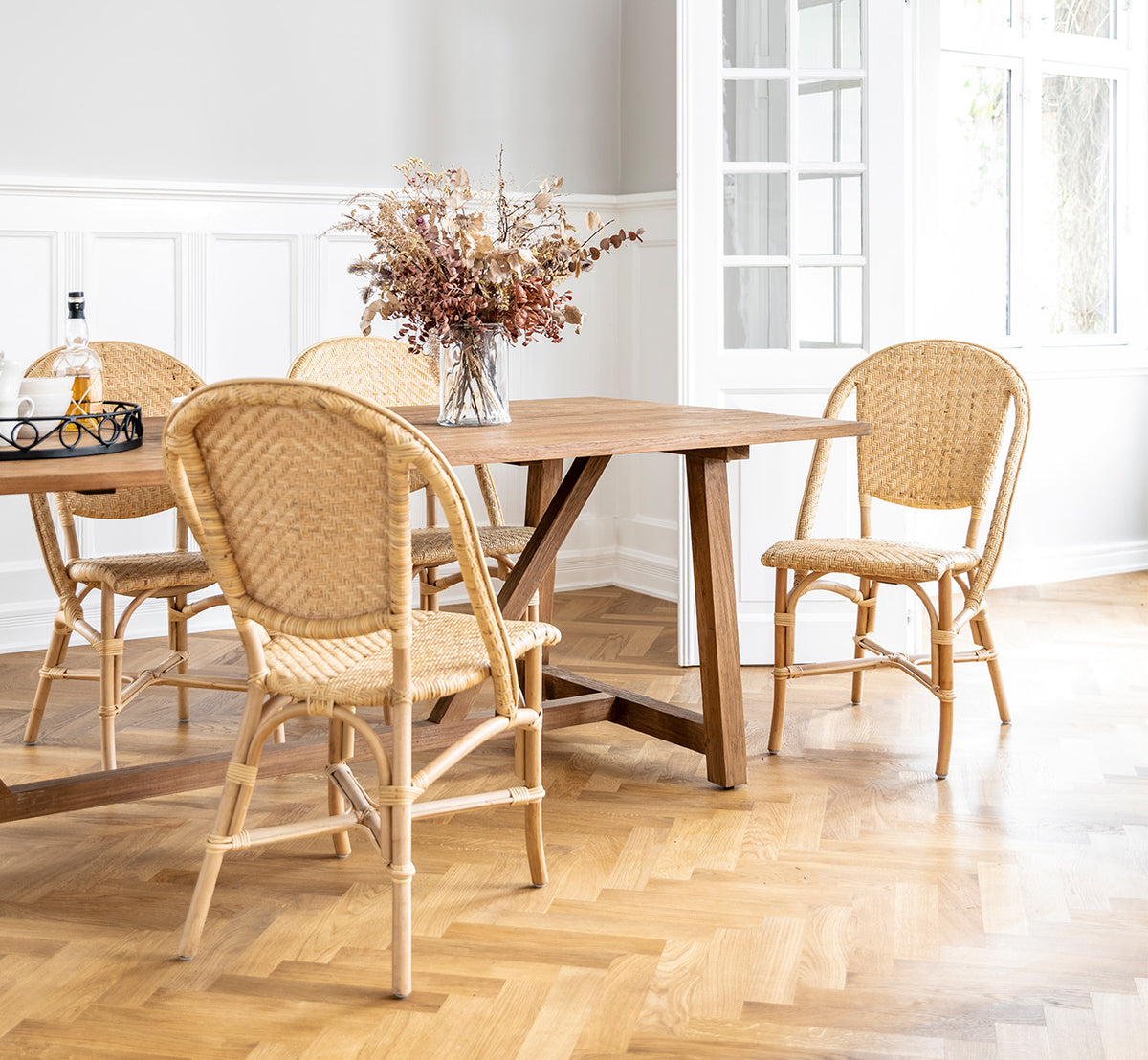 This is a lifestyle image of Alanis Dining Chair in natural rattan, in a bright interior with white panelled walls and herringbone parquet.