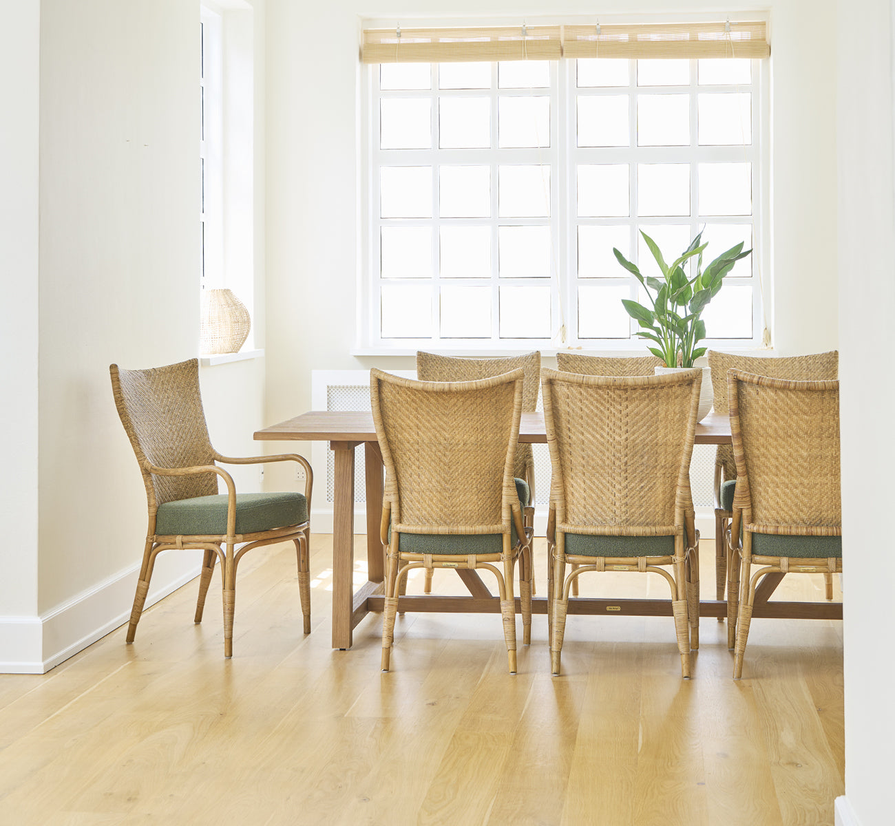 This is a lifestyle image of Melody Dining Armchair in antique rattan. Six chairs at a teak table in a bright room with oak floor.