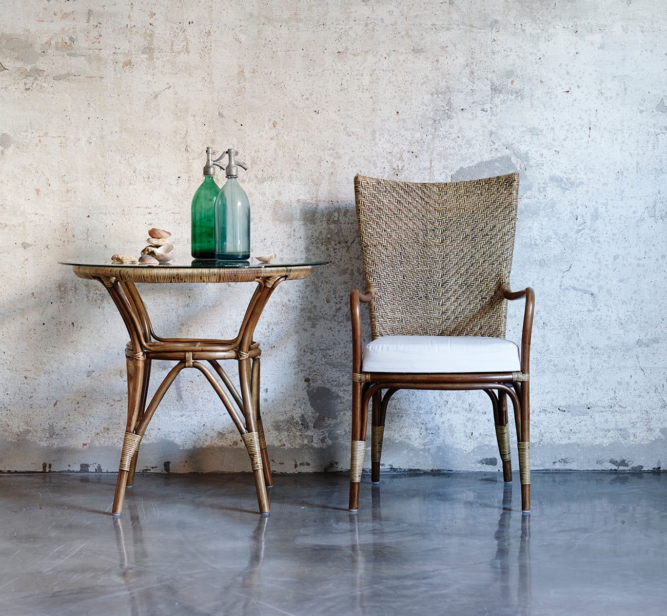 This is a lifestyle image of Melody Dining Armchair in antique rattan. Paired with a glass-top side table against a concrete wall.