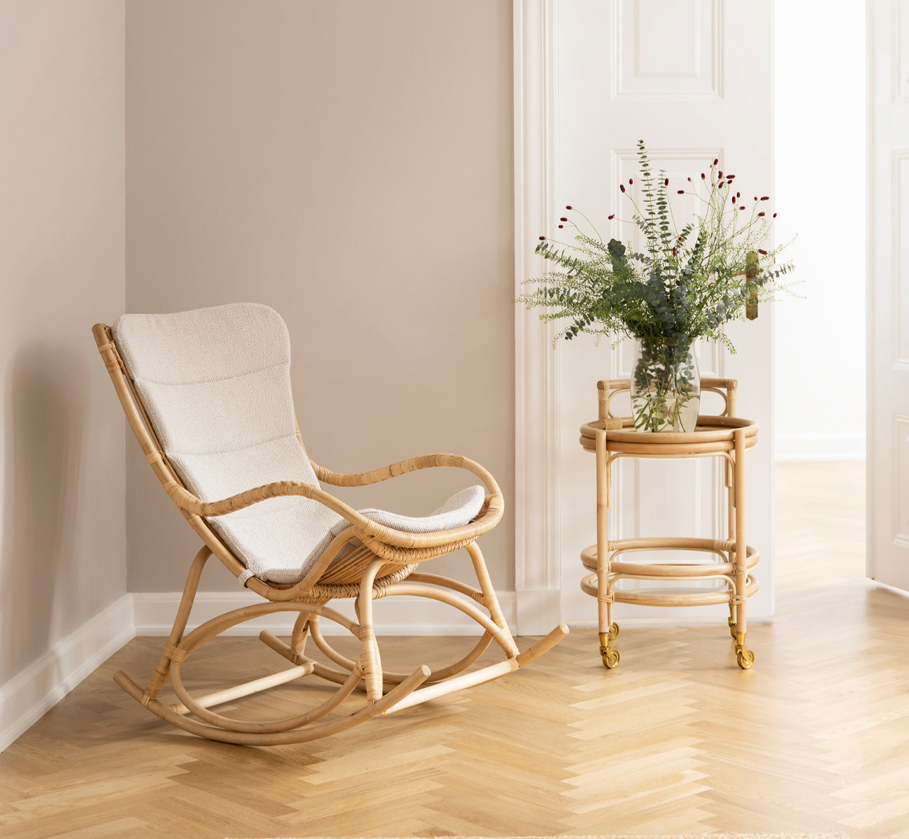 This is a lifestyle image of Monet Rocking Chair in natural rattan. In a blush-walled room with herringbone parquet and eucalyptus.