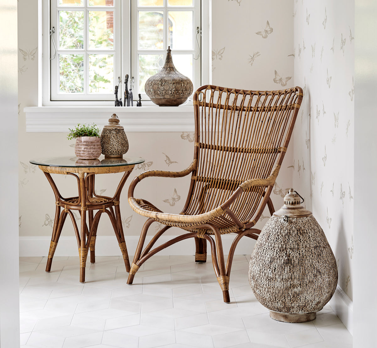 This is a lifestyle image of Monet Lounge Chair in antique rattan. With footstool in a rustic white room with dark navy textile.