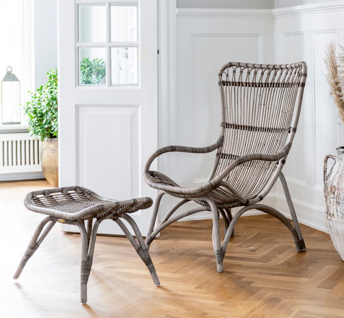 This is a lifestyle image of Monet Lounge Chair in taupe rattan. With footstool in a dark barn setting with candle and glassware.