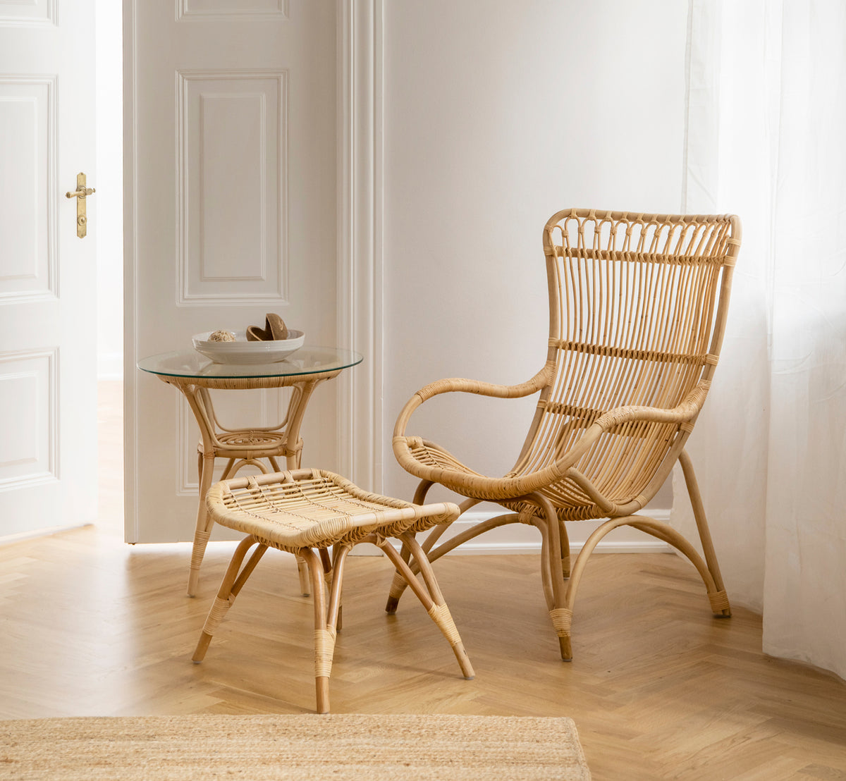 This is a lifestyle image of Monet Lounge Chair in natural rattan. With footstool and side table near a doorway on herringbone floor.