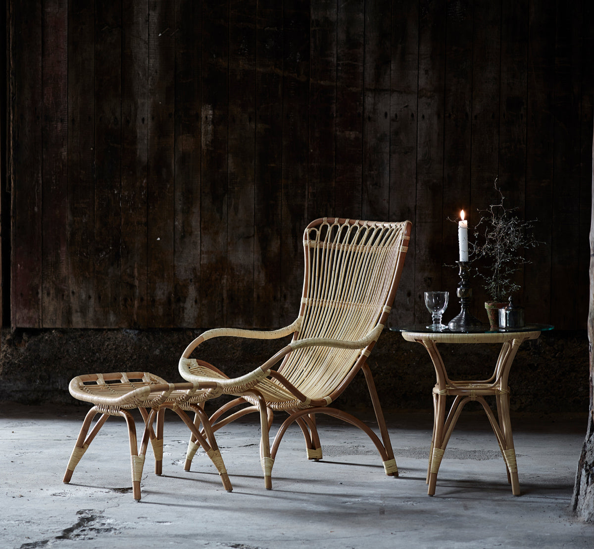 This is a lifestyle image of Monet Lounge Chair in natural rattan. Paired with a footstool and side table against a dark wood-clad wall.