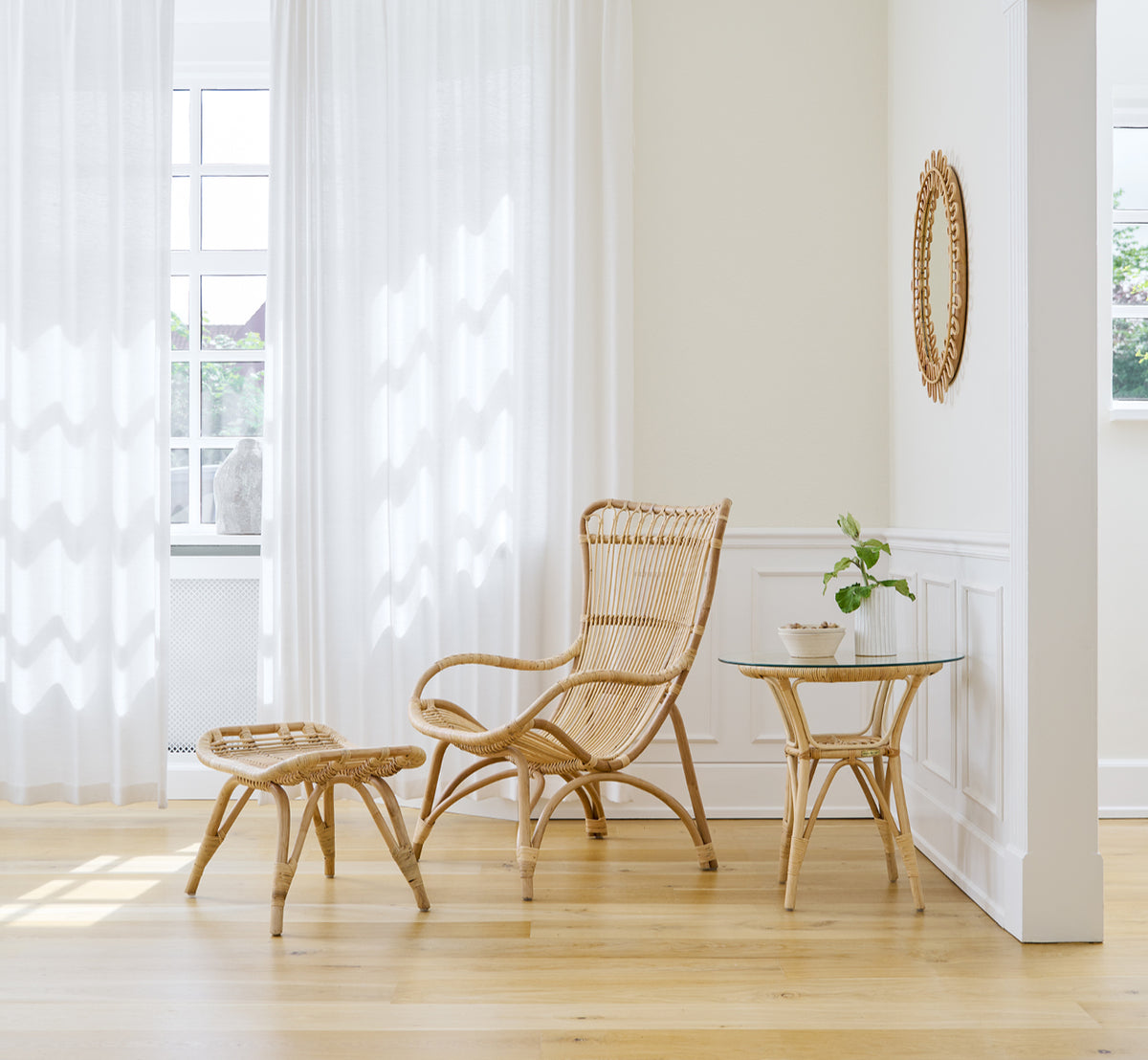 This is a lifestyle image of Monet Footstool in natural rattan. With lounge chair in a bright room with sheer curtains and oak floor.