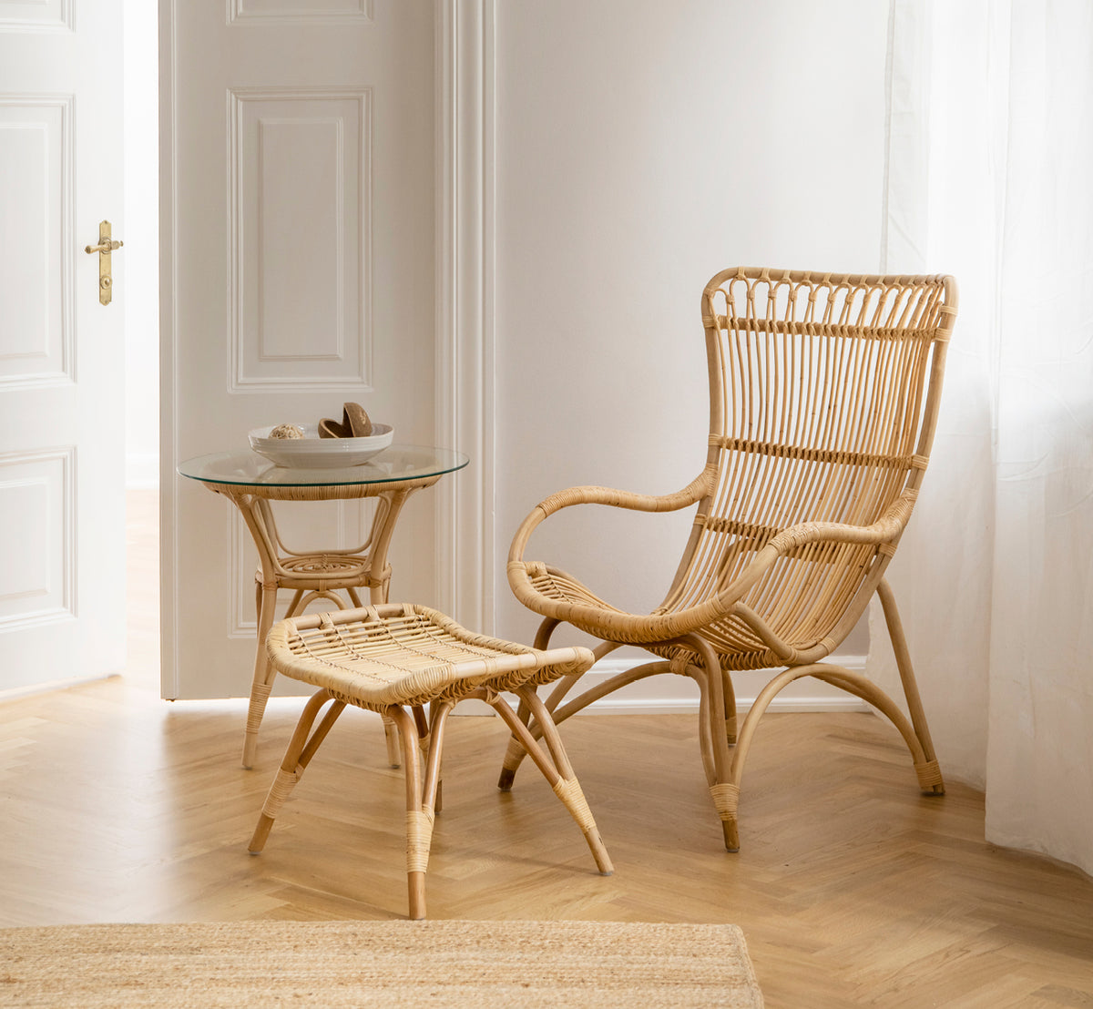 This is a lifestyle image of Monet Footstool in natural rattan. Paired with a high-back rattan chair and glass side table in a bright entry.