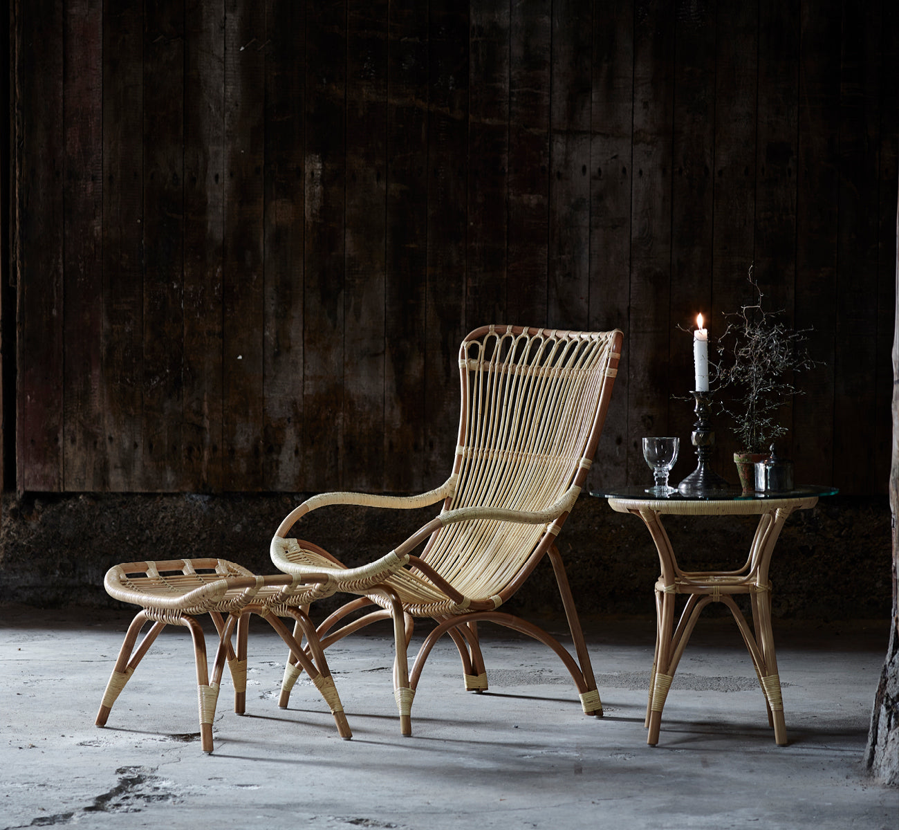 This is a lifestyle image of Monet Footstool in natural rattan. With lounge chair in a dark rustic barn setting with glassware.