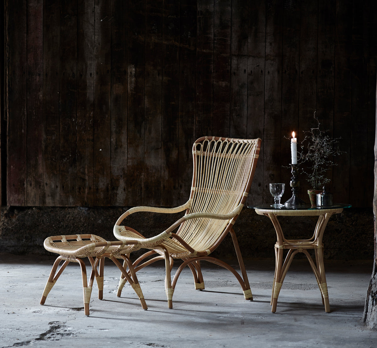 This is a lifestyle image of Monet Footstool in natural rattan. With lounge chair in a dark rustic barn setting with glassware.