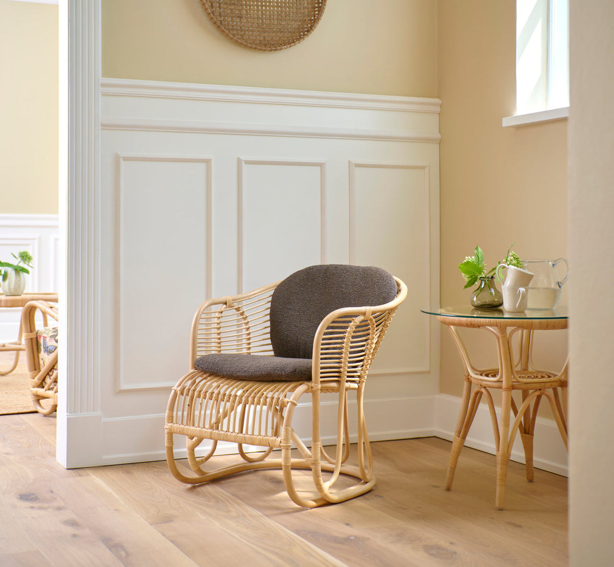 This is a lifestyle image of Tony Side Table in natural rattan. Beside an armchair in a bright paneled room.