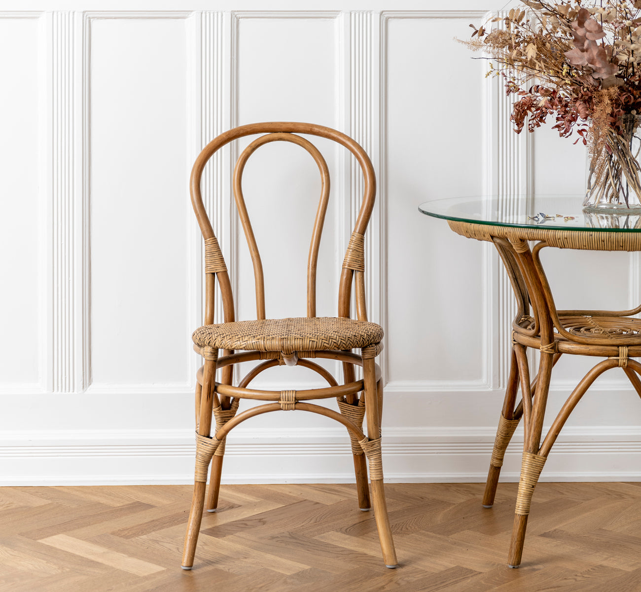 This is a lifestyle image of Tony Café Table in antique rattan. Paired with a dining chair beside dried flowers in a white paneled interior.