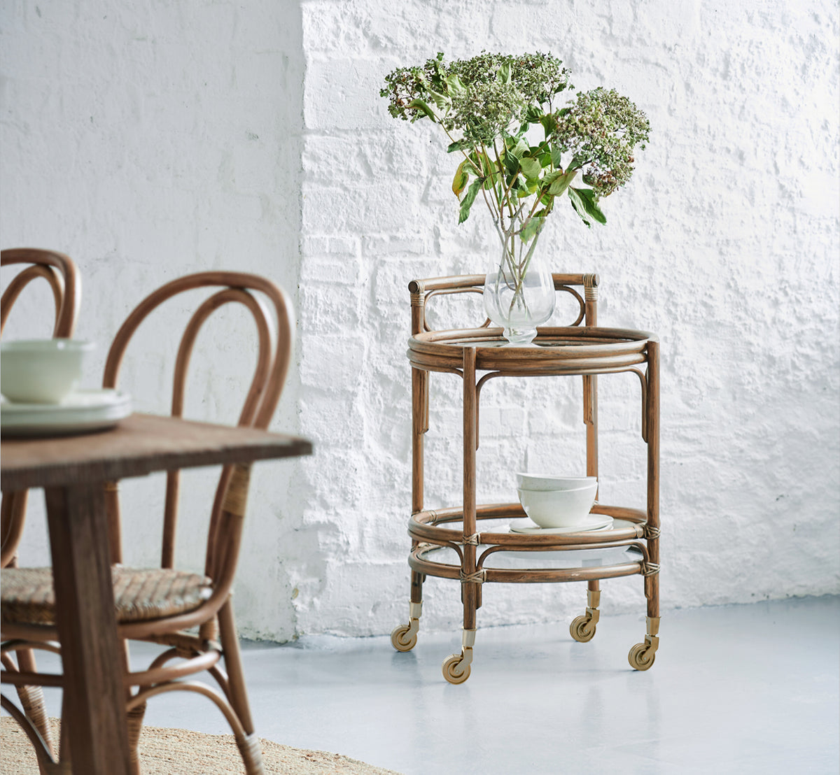 This is a lifestyle image of Romeo Trolley in antique rattan. Styled with hydrangeas and ceramics against a white brick wall.