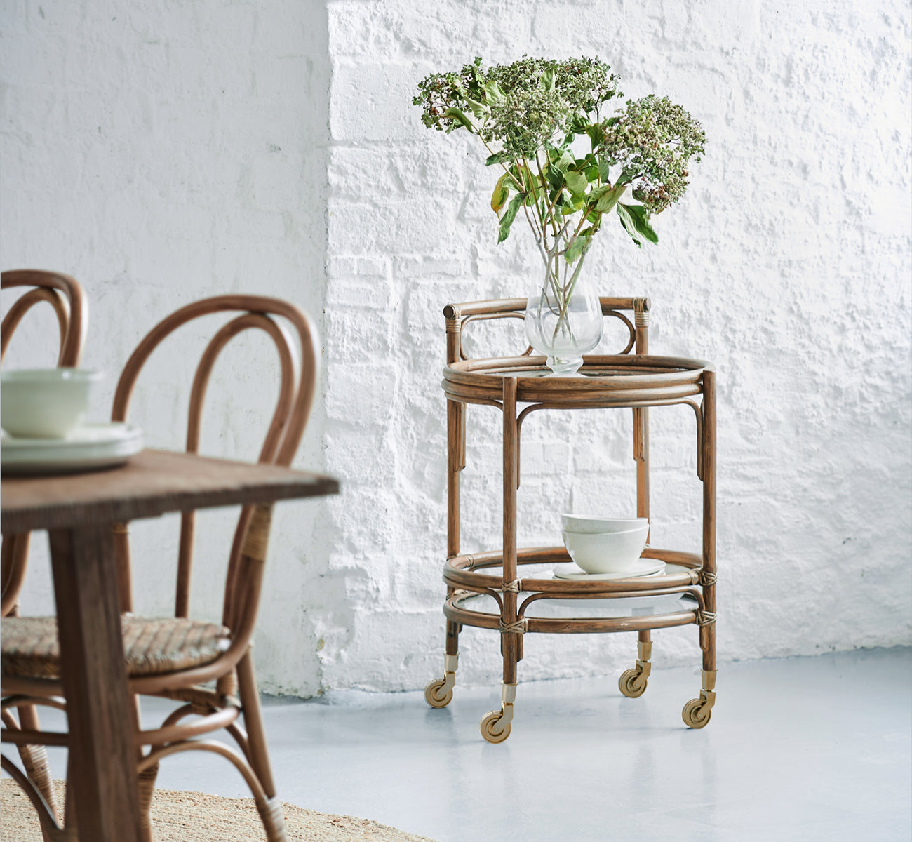 This is a lifestyle image of Romeo Trolley in antique rattan. Styled with hydrangeas and ceramics against a white brick wall.
