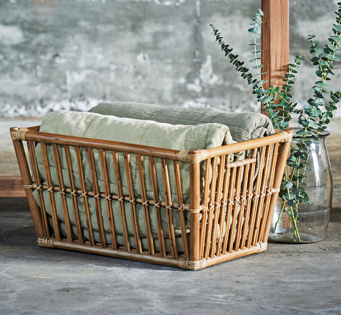 This is a lifestyle image of Marche Basket in antique rattan. Styled with linen blankets and eucalyptus in a concrete-walled room.