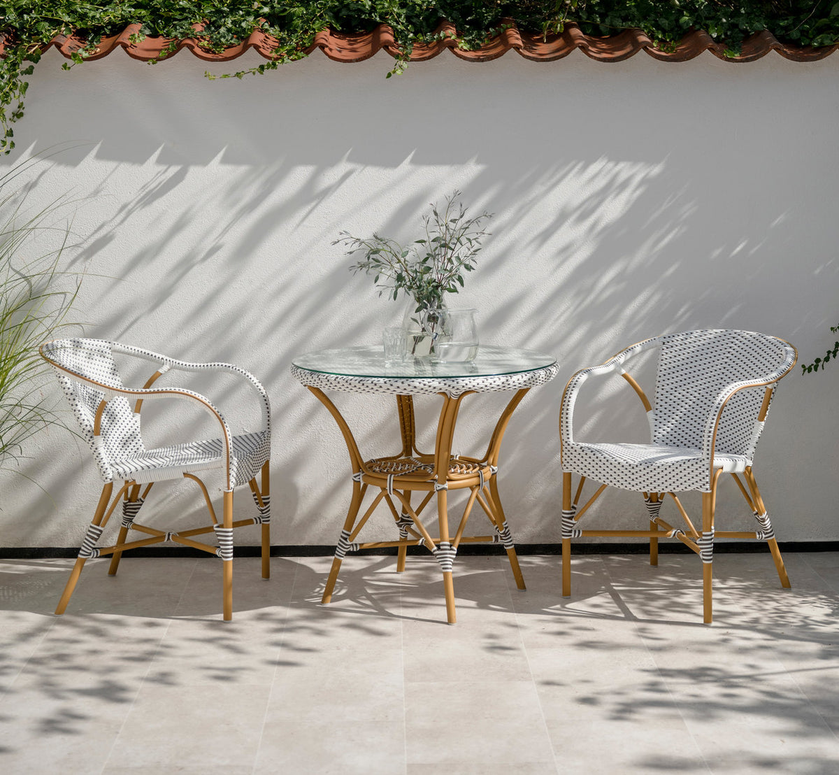 This is a lifestyle image of Madeleine Exterior Dining Chair in almond aluminium frame white w cappuccino dot artfibre. At a glass table on a terrace.