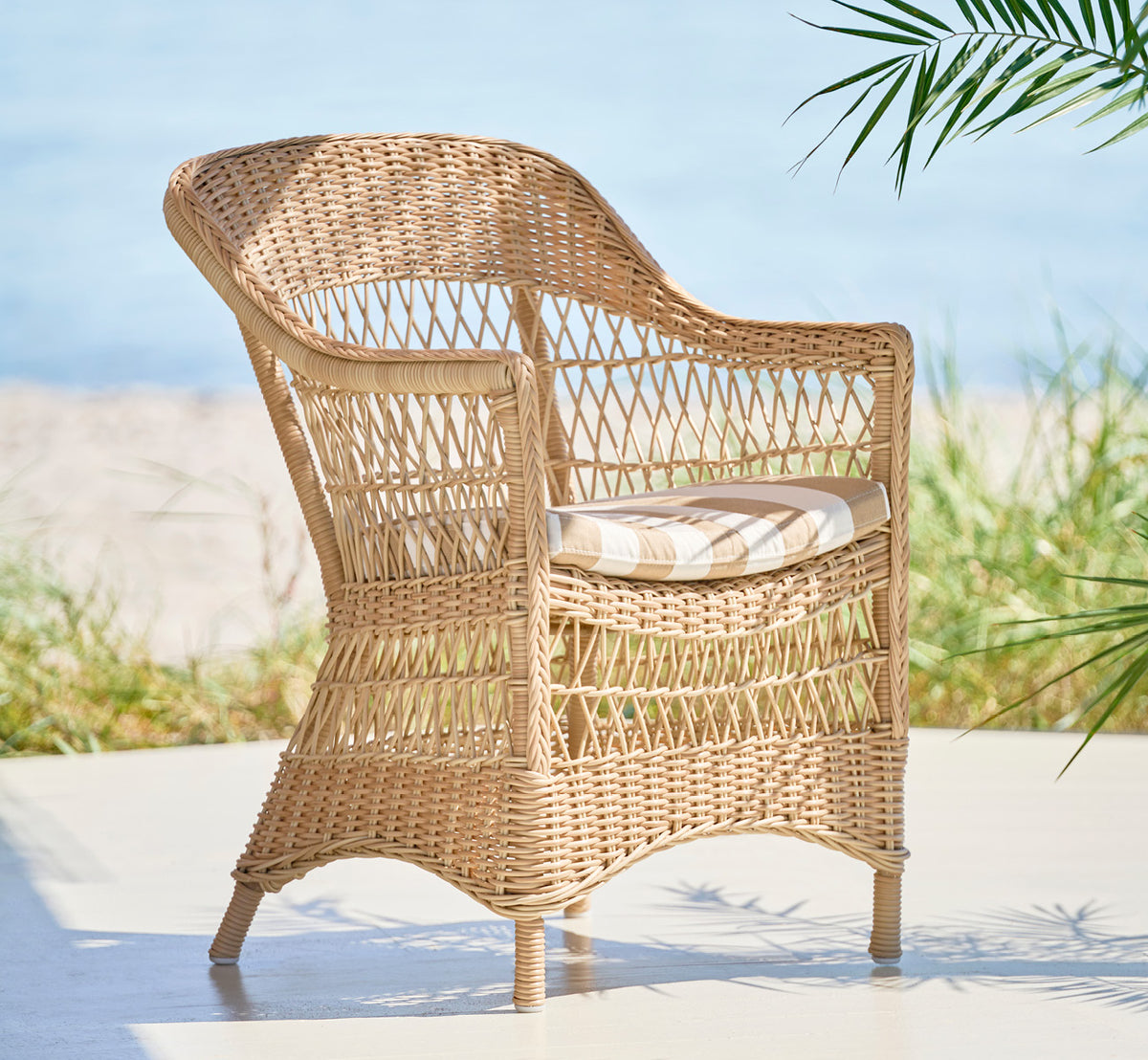 This is a lifestyle image of Charlot Chair in natural ArtFibre Aluminium, on a sun-drenched coastal terrace with sea views and a white pergola.