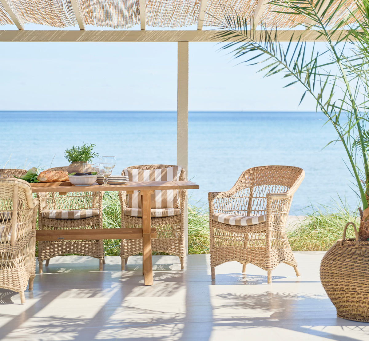 This is a lifestyle image of Charlot Chair in natural ArtFibre Aluminium, on a sun-drenched coastal terrace with sea views and a white pergola.