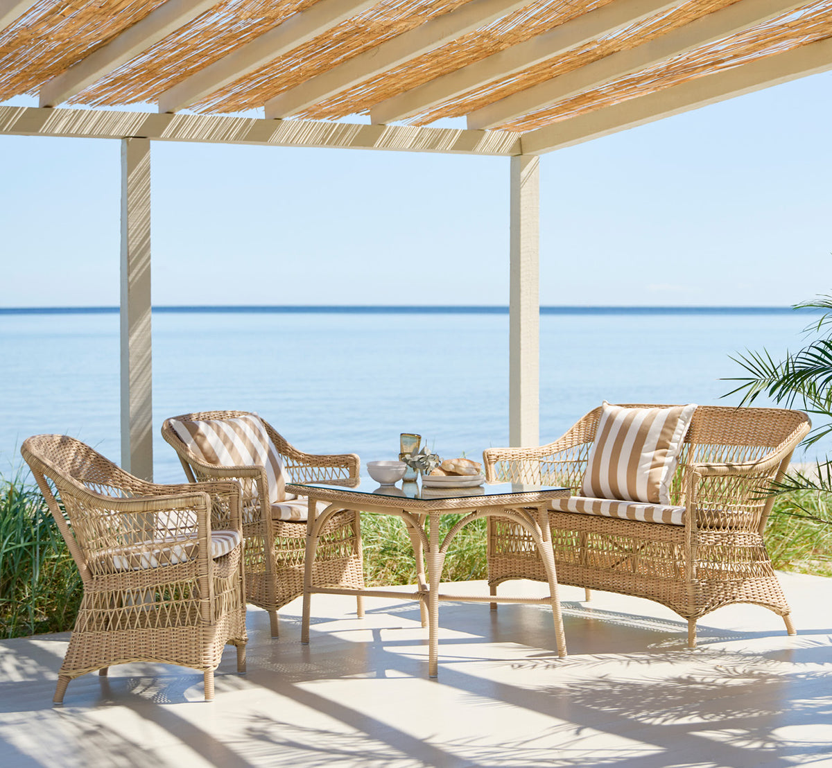 This is a lifestyle image of Charlot Chair in natural ArtFibre Aluminium, on a sun-drenched coastal terrace with sea views and a white pergola.