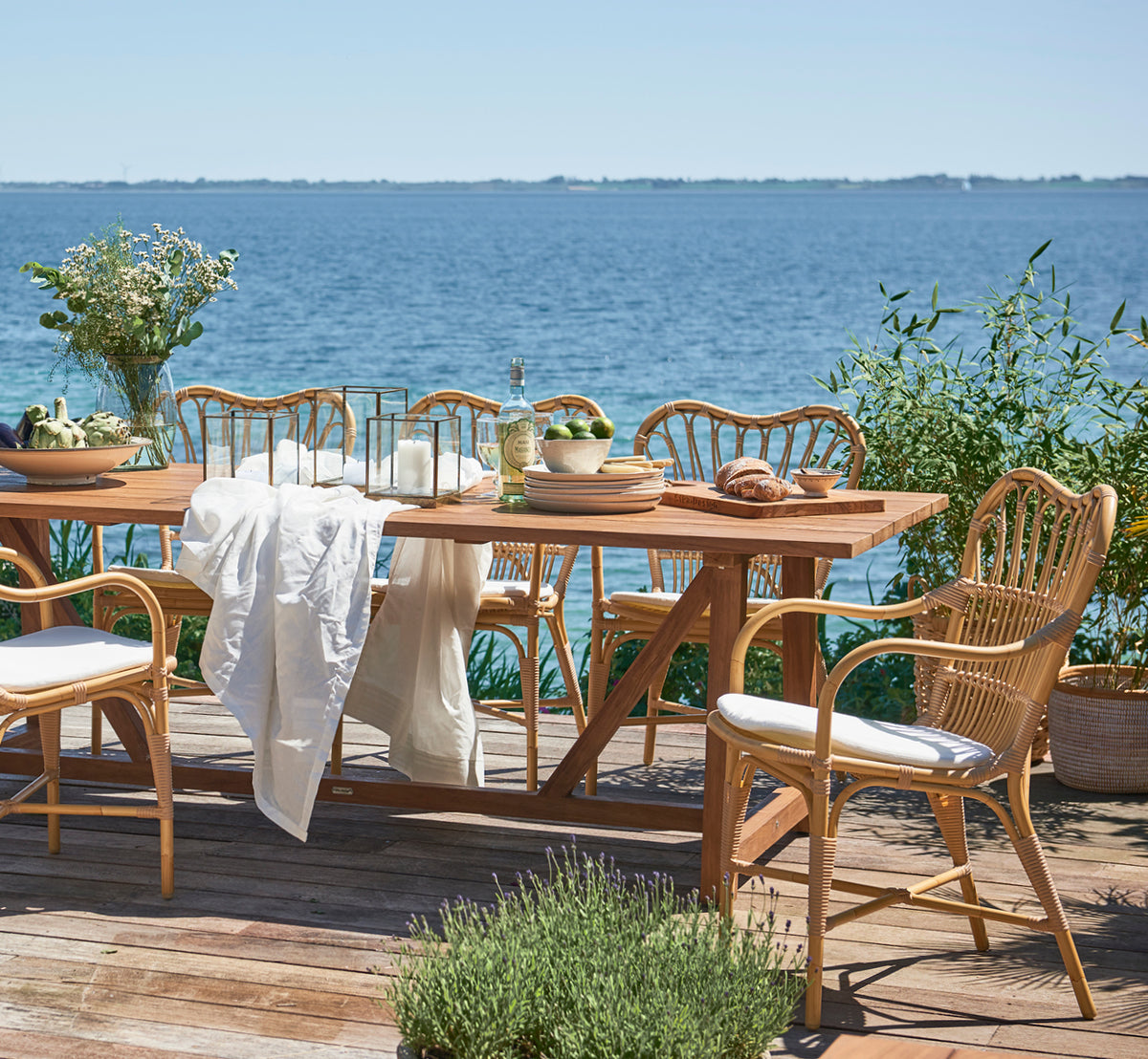 This is a lifestyle image of George Exterior Table 240 in outdoor Teak, on an outdoor garden patio with a white house facade and greenery.