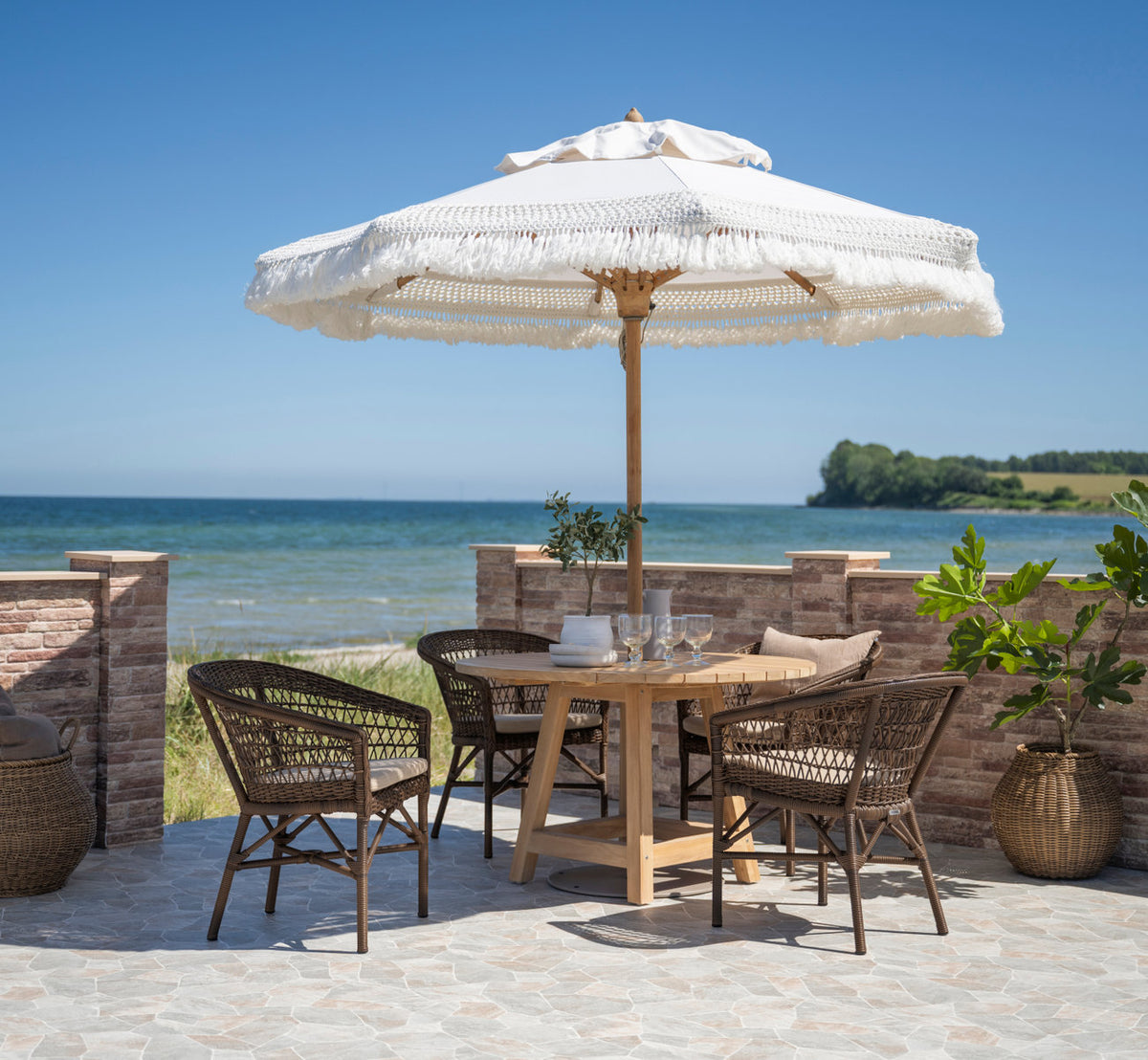 This is a lifestyle image of George Teak Table Ø120 cm in teak outdoor. Dining under a fringed parasol by the sea.