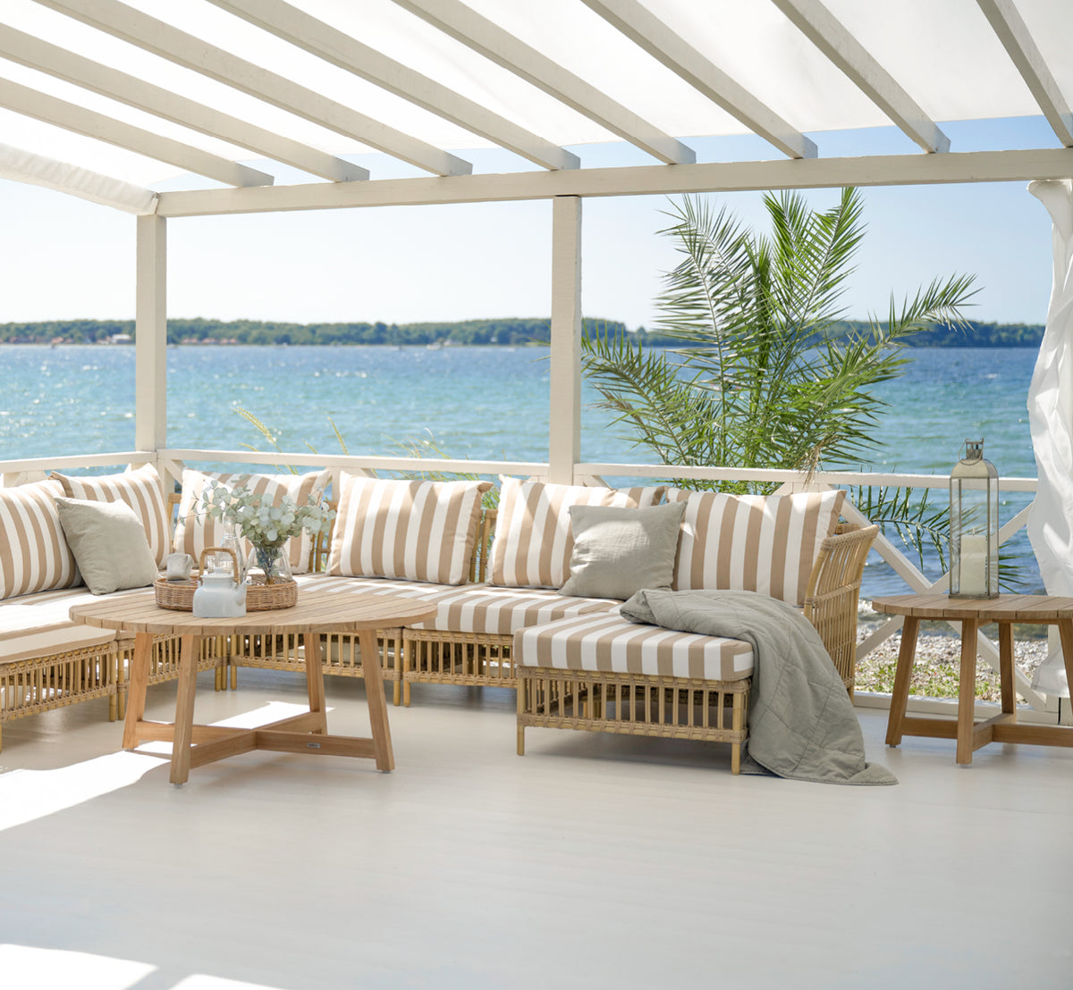 This is a lifestyle image of Victor Teak Lounge Table Ø120 cm in teak outdoor. Placed between outdoor sofas with striped cushions overlooking the sea.