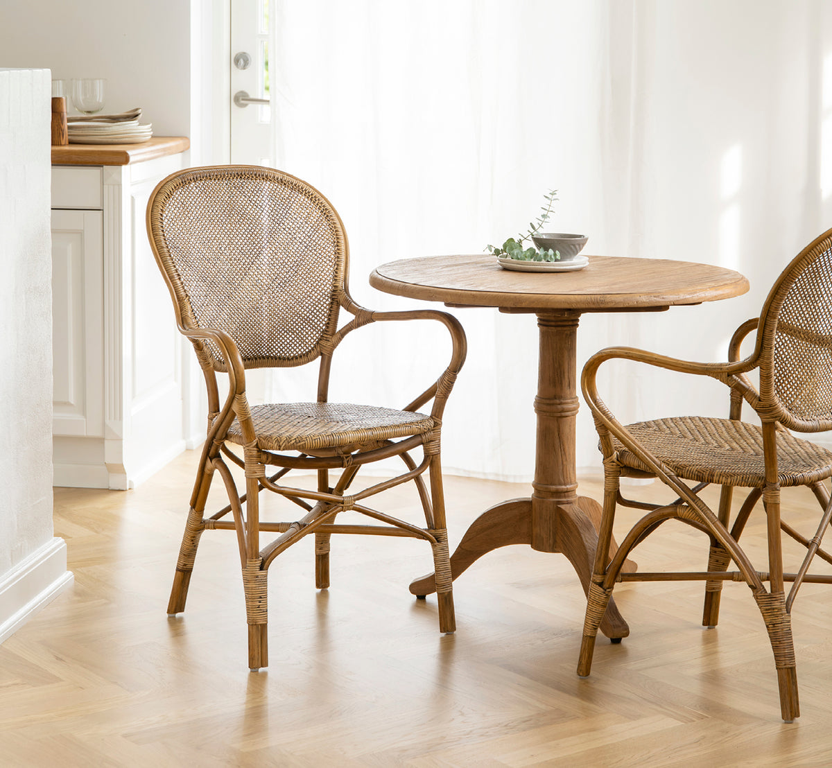 This is a lifestyle image of Michel Teak Dining Table Ø80 cm in teak indoor. Paired with rattan armchairs in a light, airy café-style interior.
