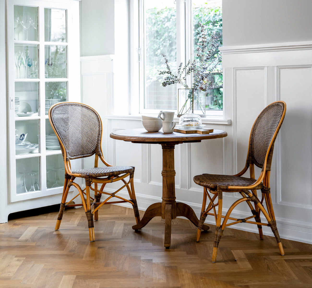 This is a lifestyle image of Michel Teak Dining Table Ø80 cm in teak indoor. Set in a warm rustic dining space with rattan chairs and soft lighting.
