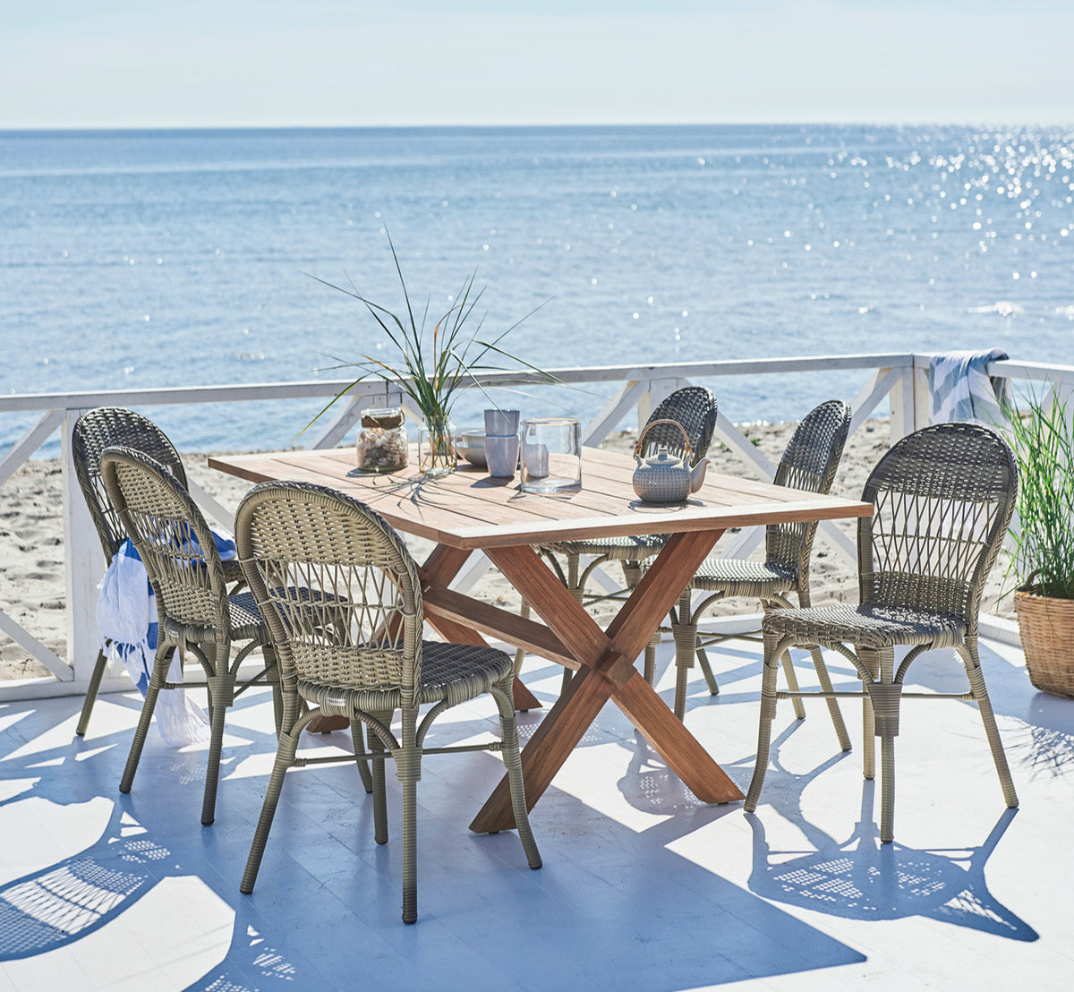This is a lifestyle image of Colonial Table 160 in outdoor Teak, on a beachside terrace with white wooden decking and sea views.