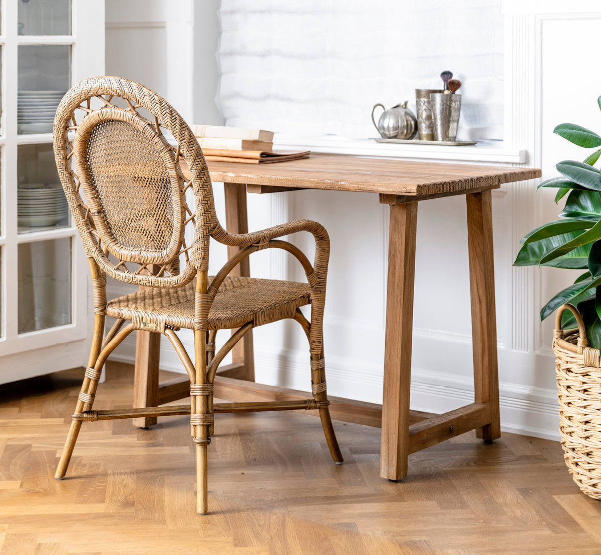 This is a lifestyle image of Lucas Teak Desk in teak indoor. Paired with a rattan chair and green plant in a bright interior with herringbone floor.
