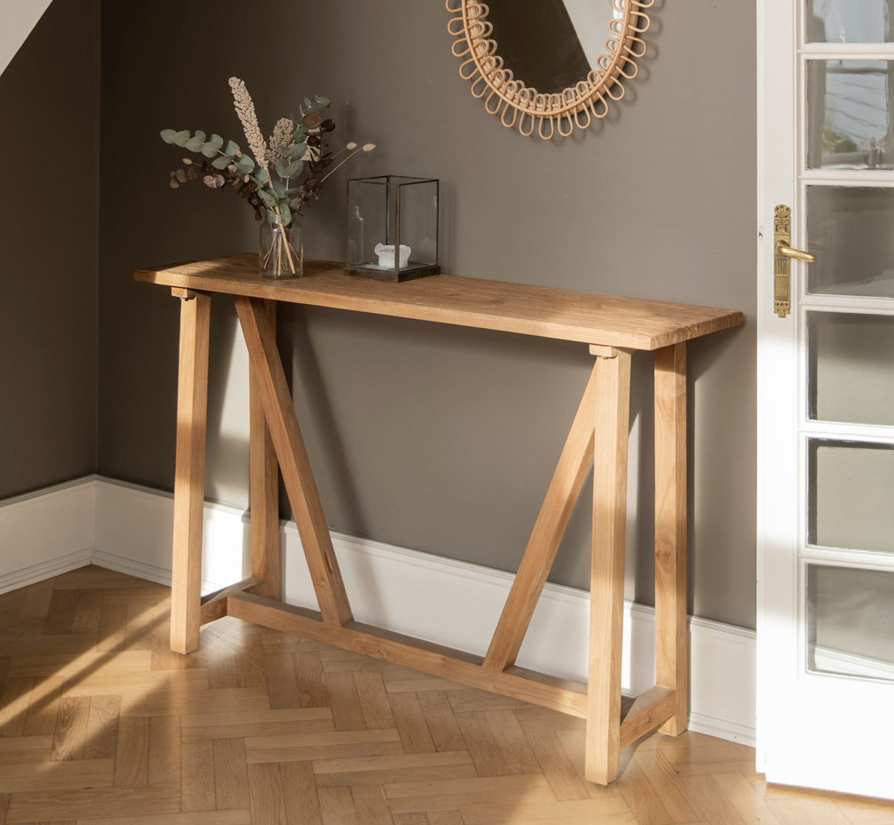This is a lifestyle image of Lucas Teak Console in teak indoor. Against a dark gray wall with a rattan mirror above and eucalyptus in a vase.