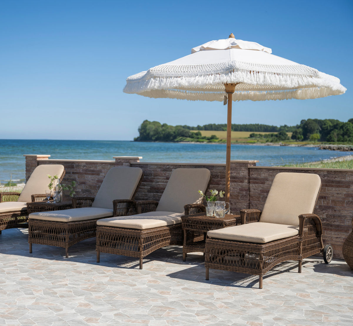 This is a lifestyle image of Susy Exterior Side Table in brown artfibre aluminum. Row of sunbeds under a fringed parasol by the sea.