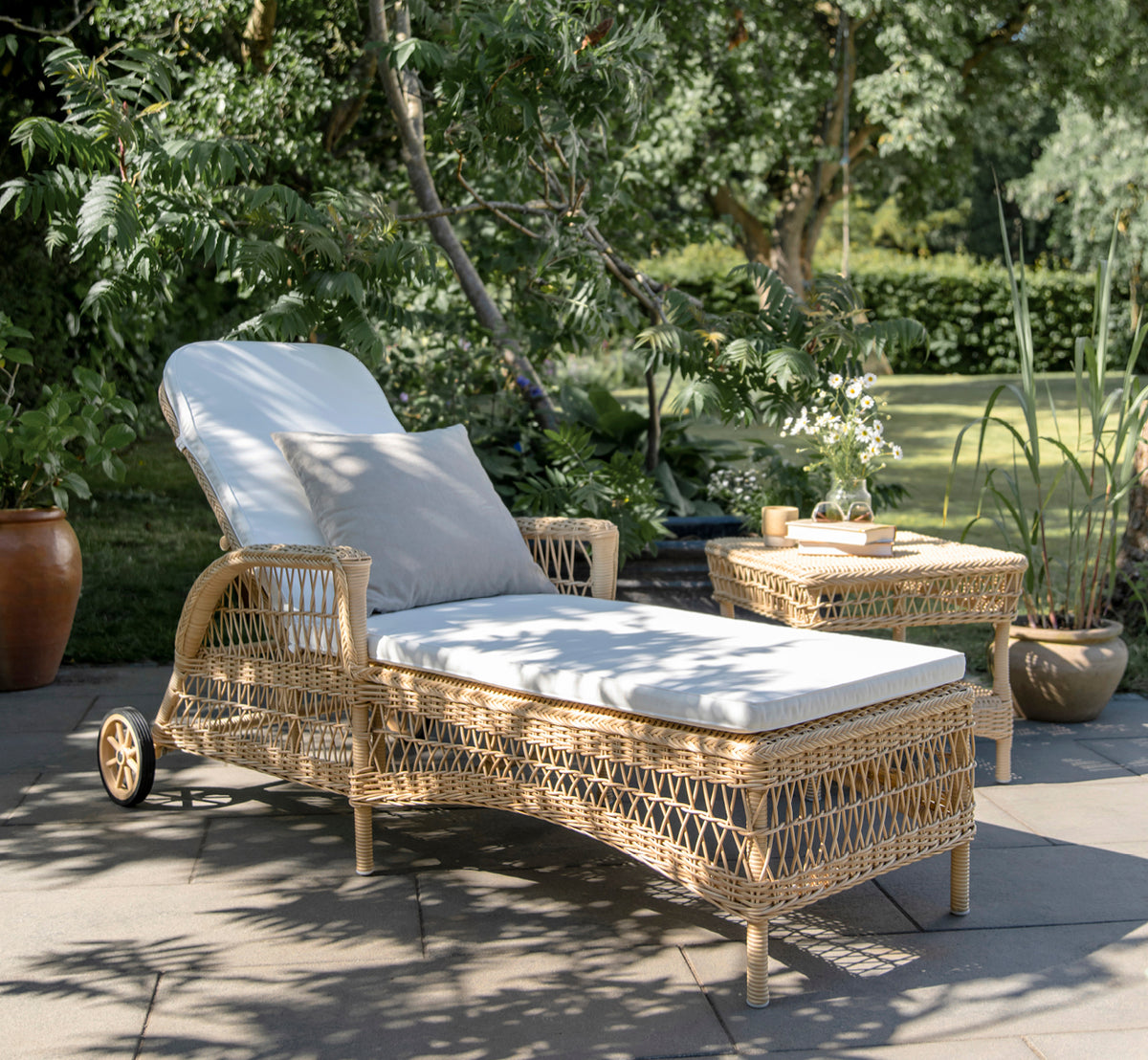 This is a lifestyle image of Daisy Sunbed in natural ArtFibre Aluminium, on a garden terrace surrounded by lush trees and potted plants.