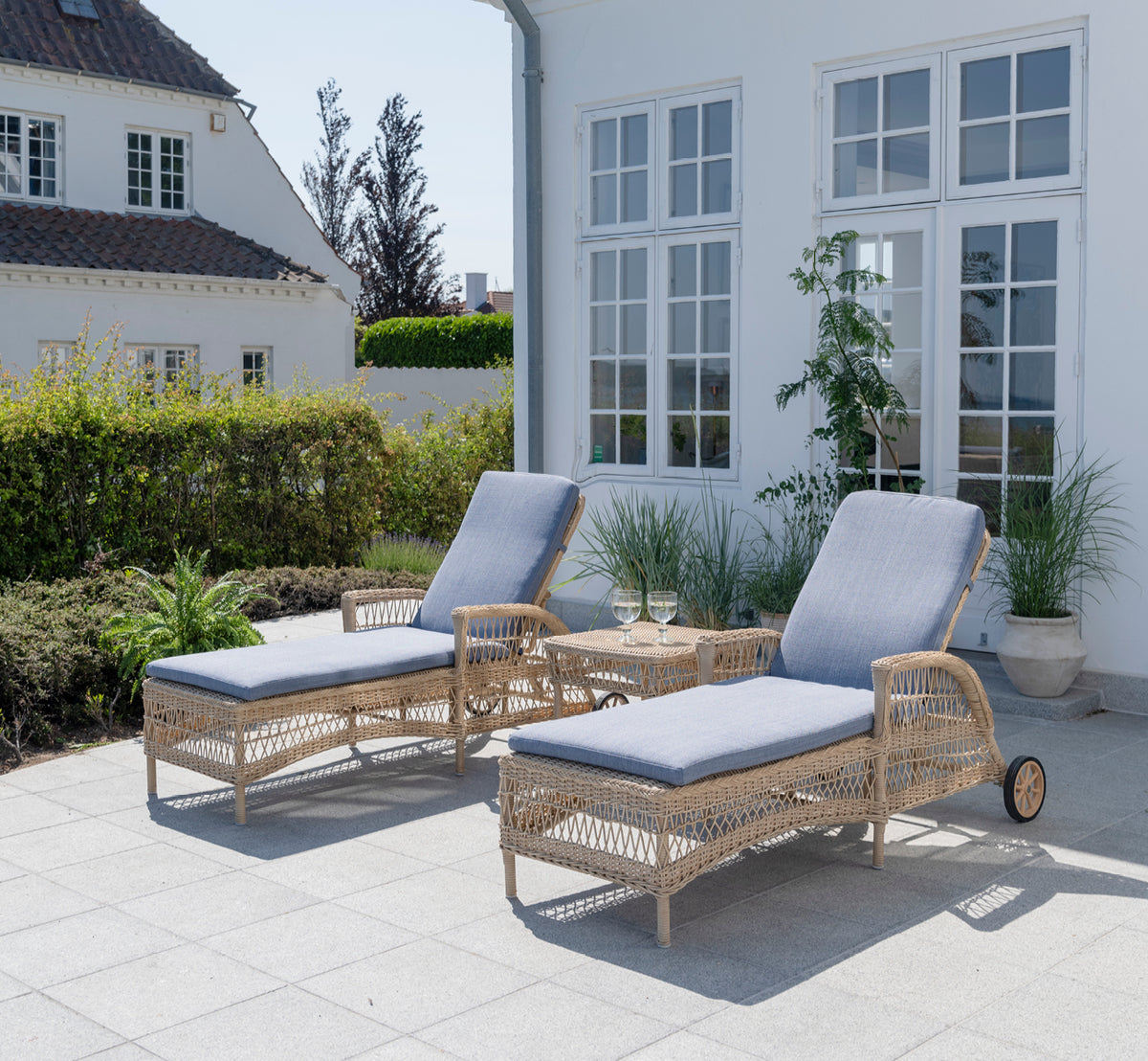 This is a lifestyle image of Daisy Sunbed in natural ArtFibre Aluminium, on a garden terrace surrounded by lush trees and potted plants.