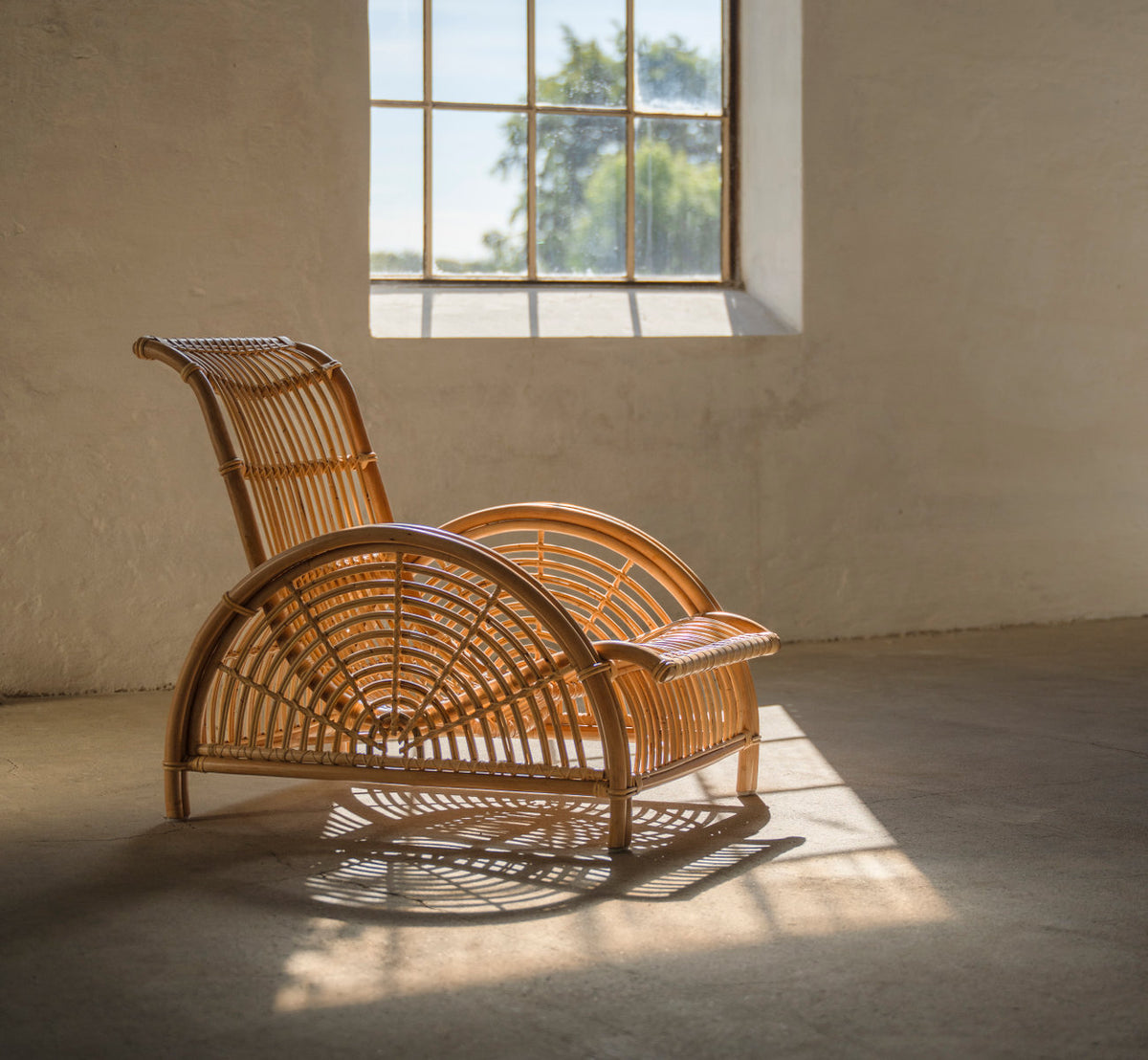This is a lifestyle image of Paris Lounge Chair in polished natural rattan. In warm sunlight by a window in a rustic room.