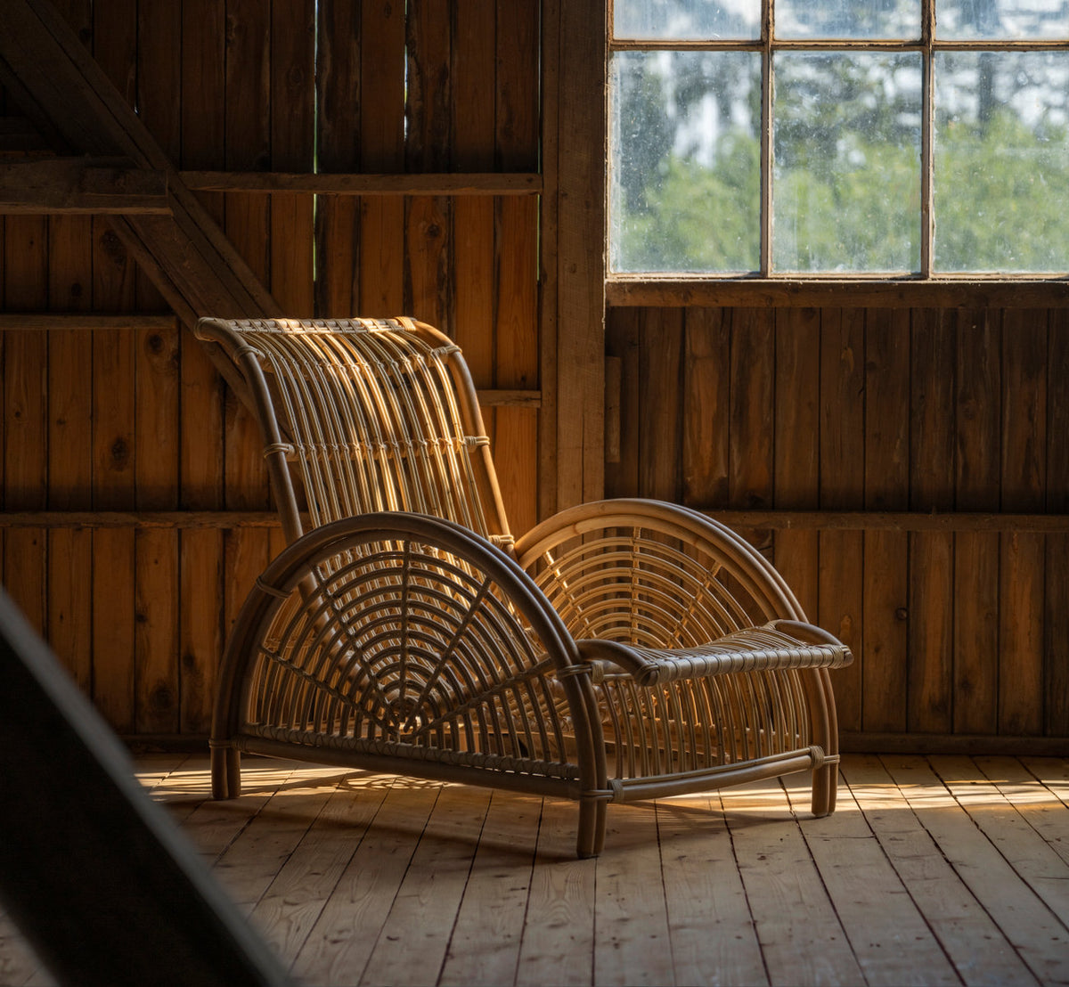 This is a lifestyle image of Paris Lounge Chair in polished natural rattan. In a wooden barn with warm afternoon light.