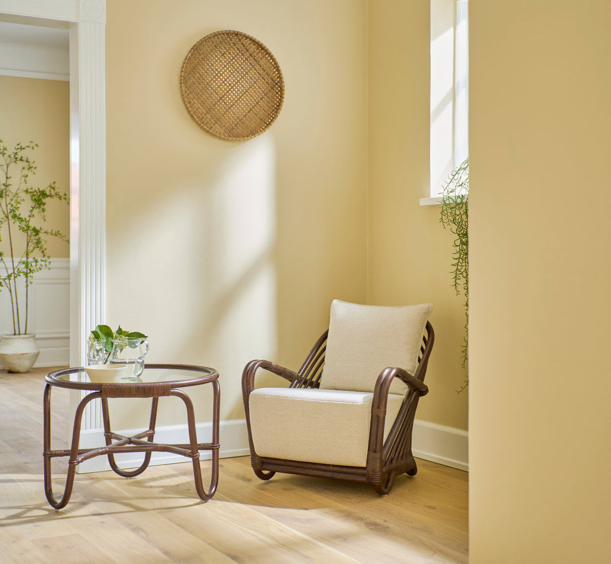 This is a lifestyle image of Charlottenborg Lounge Chair in brown rattan. With glass side table in a warm yellow corner.
