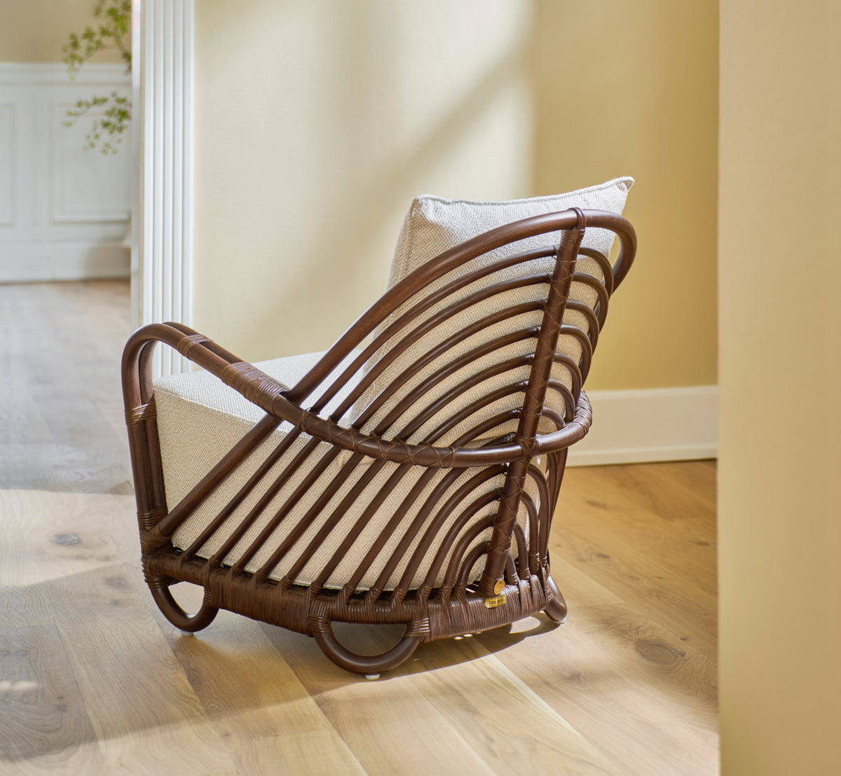 This is a lifestyle image of Charlottenborg Lounge Chair in brown rattan. With cream cushion on an oak floor by a doorway.