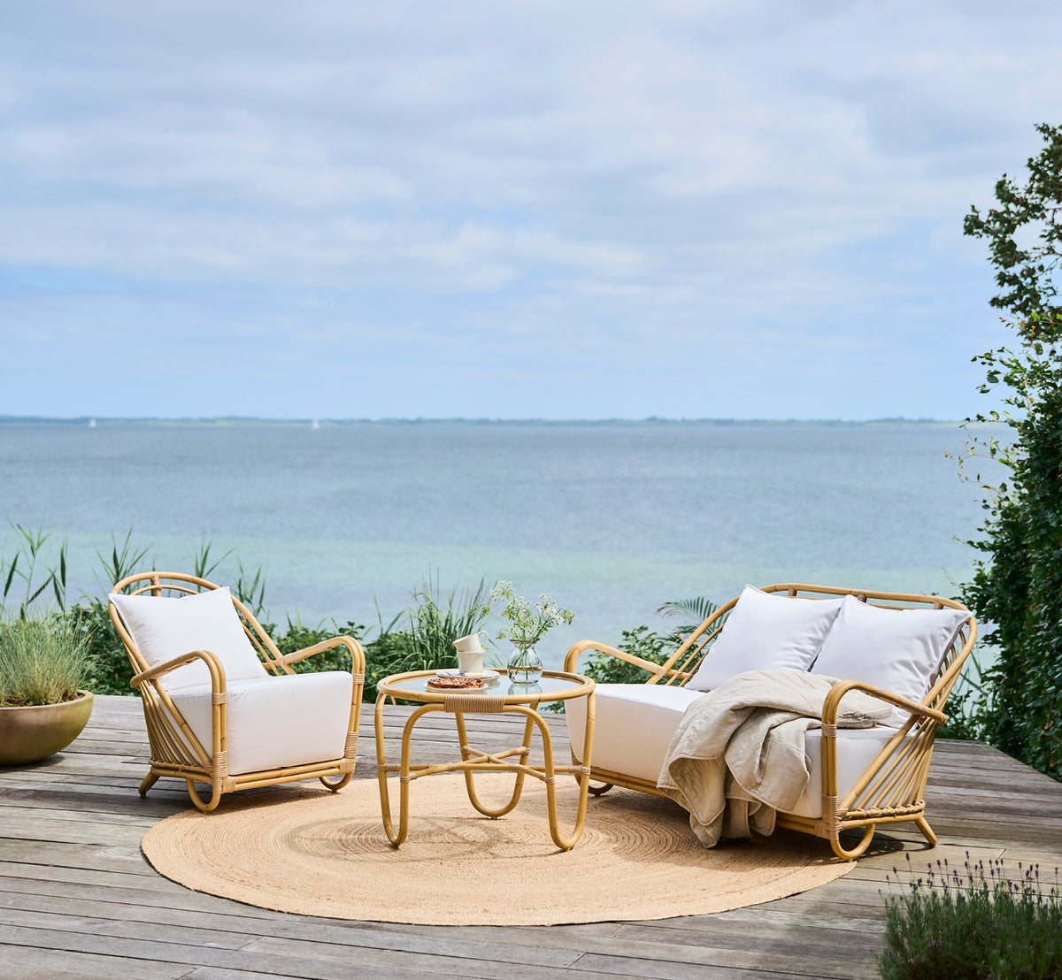 This is a lifestyle image of Charlottenborg Exterior Coffee Table in natural Aluminium ArtFibre, on an outdoor wooden deck with coastal greenery and.