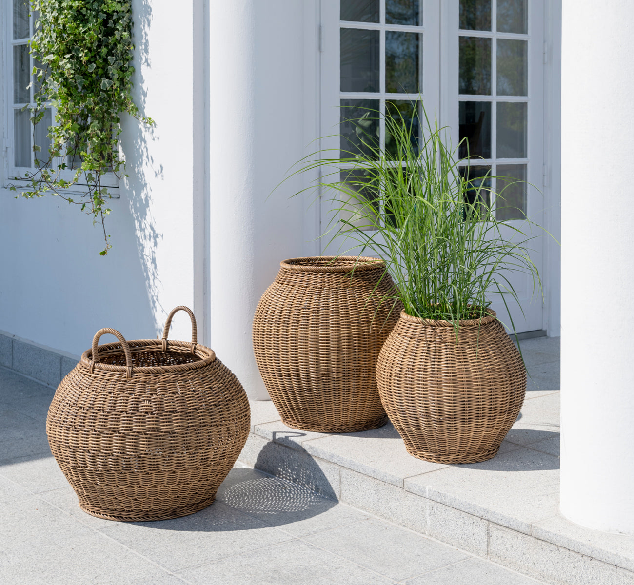 This is a lifestyle image of Smilla Basket in chestnut core weave ArtFibre Aluminium, outdoors on a sun-lit patio with a white wall and tile flooring.