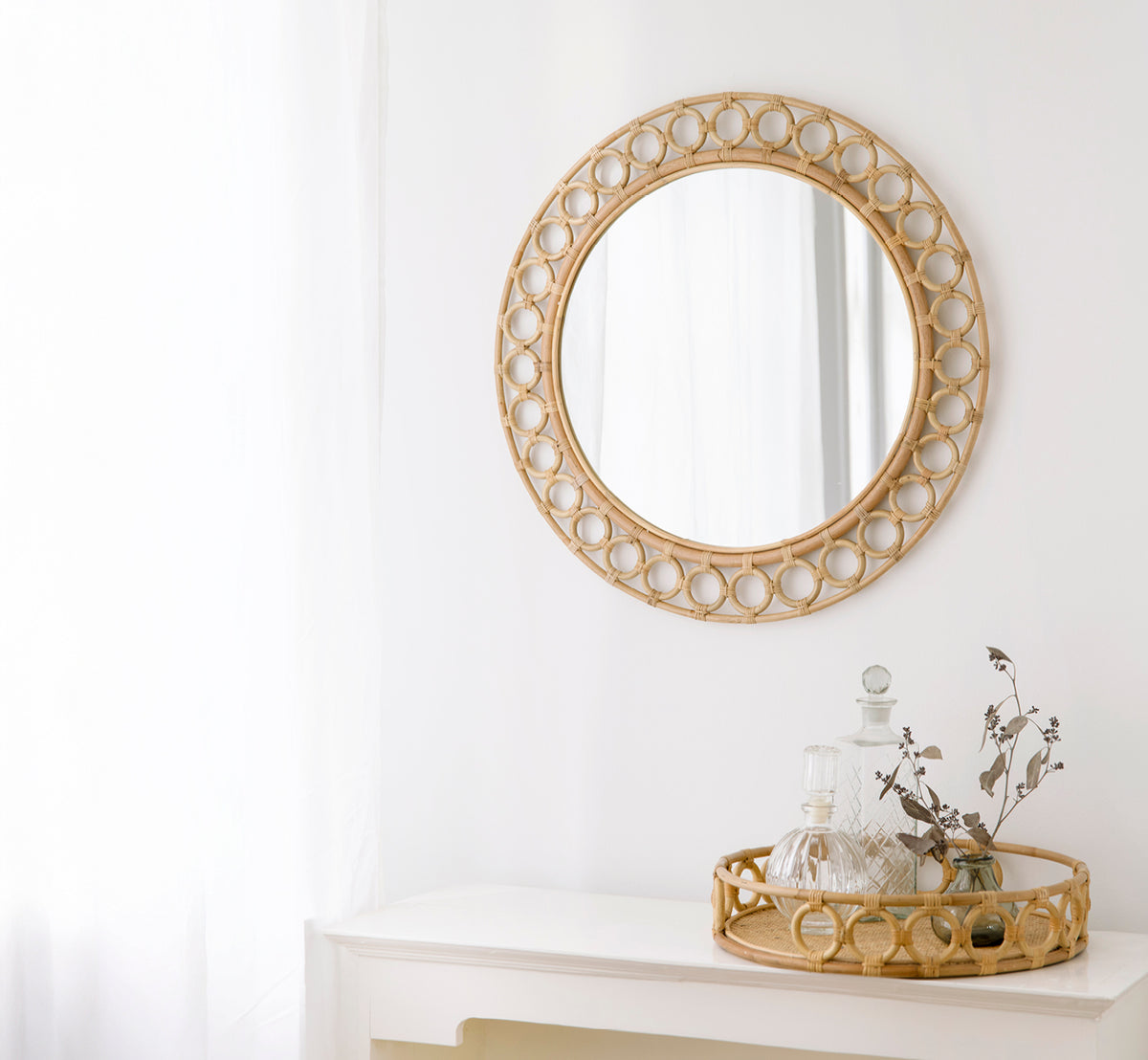 This is a lifestyle image of Mira Mirror in natural rattan. Mounted above a white console styled with decanters and dried branches.