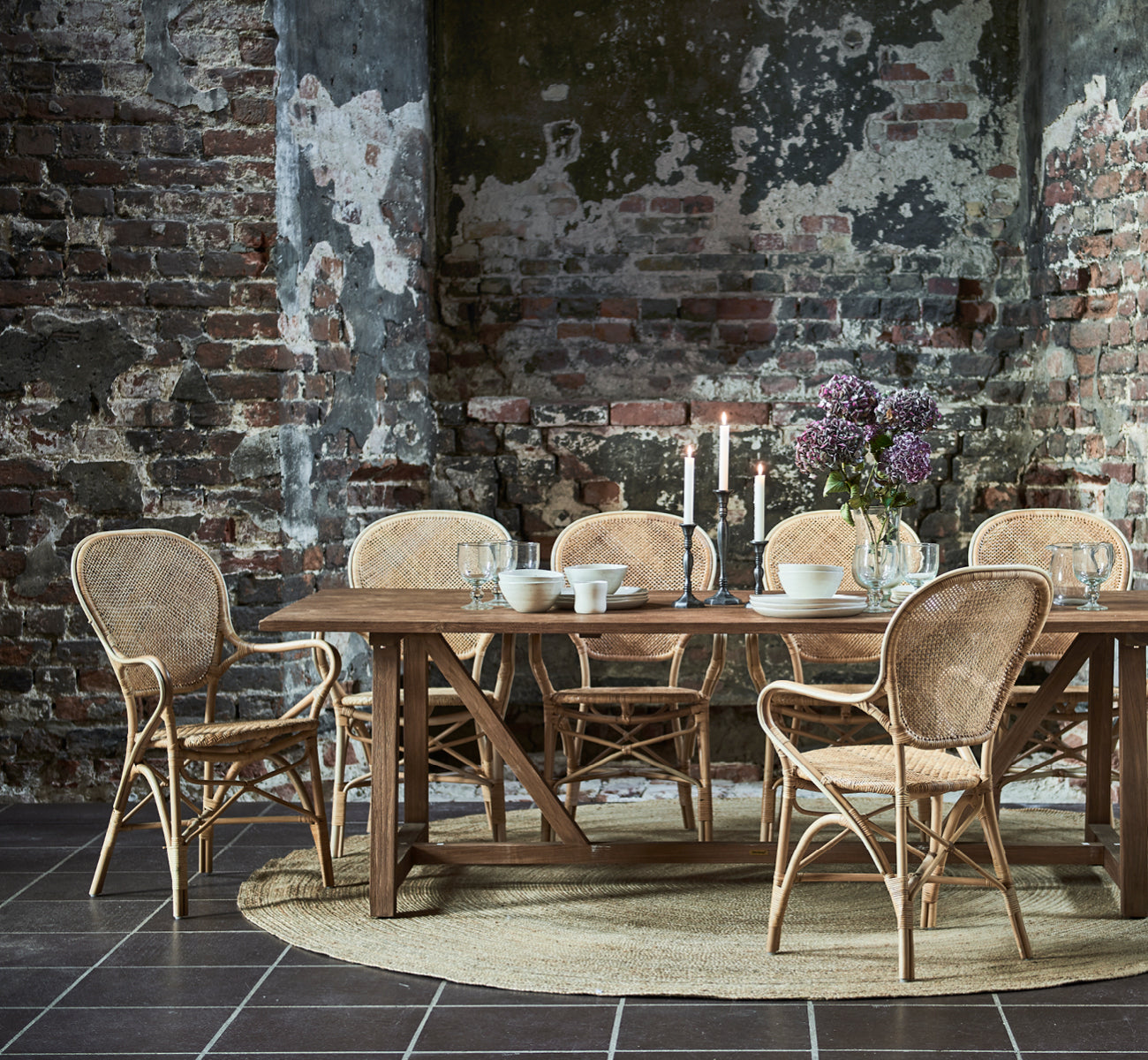 This is a lifestyle image of Jute Carpet Ø280 in natural jute. Under a dining table with rattan chairs in a rustic space with exposed brick walls.