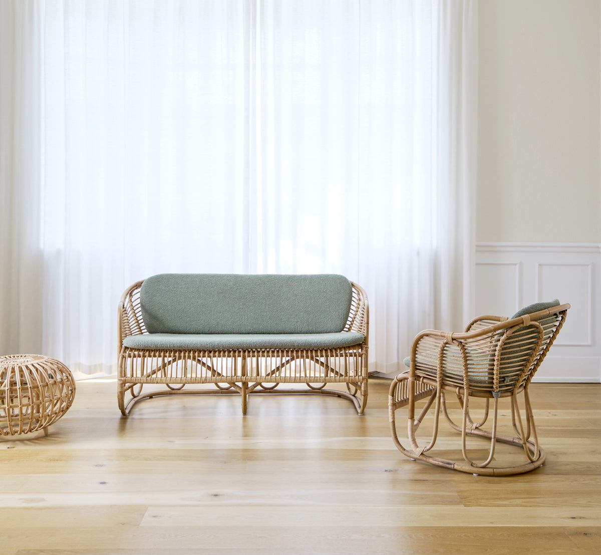This is a lifestyle image of Swing Sofa in polished natural rattan. Placed on a rug beside a fiddle-leaf plant on parquet flooring.