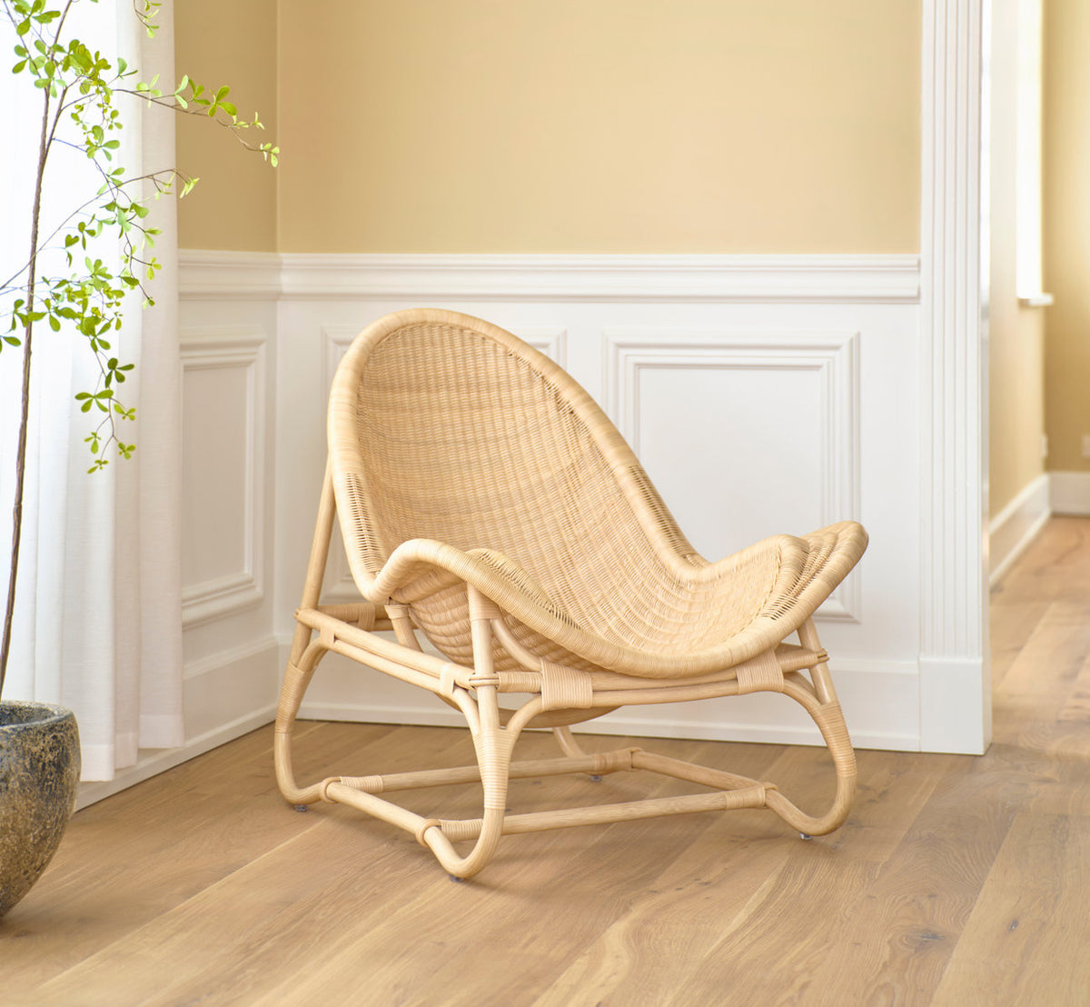This is a lifestyle image of Pacifique Lounge Chair in natural core weave rattan frame. By white panels and a potted plant.