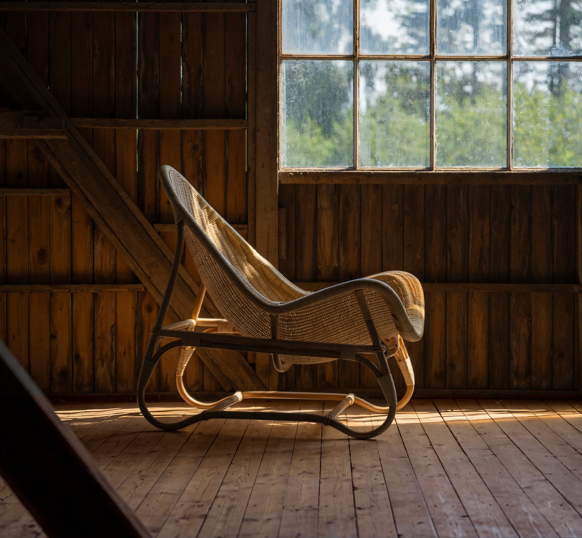 This is a lifestyle image of Pacifique Lounge Chair in natural core weave rattan frame. In a wooden barn with warm window light.