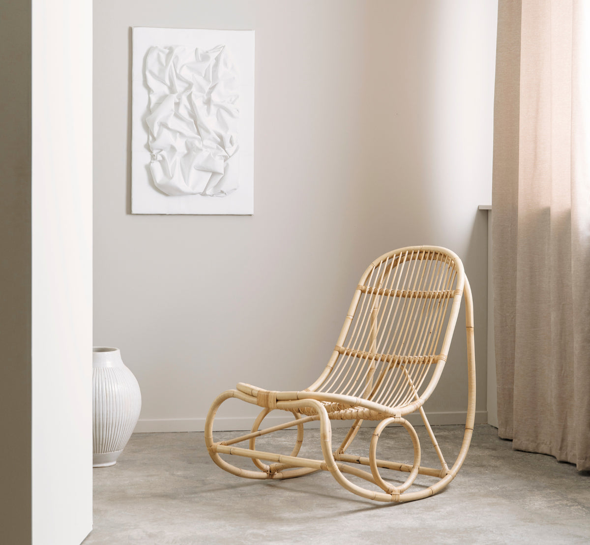 This is a lifestyle image of Nanny Rocking Chair in polished natural rattan. In a gallery setting with other rattan pieces on plinths.