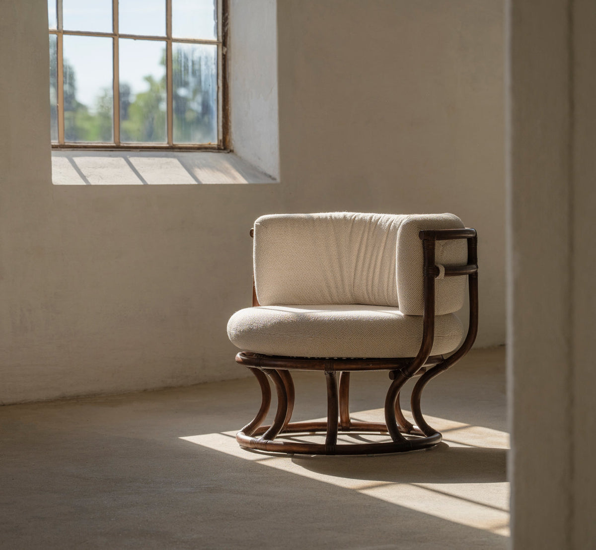 This is a lifestyle image of Coco Lounge Chair in brown rattan. With cream cushions by a sunlit window.